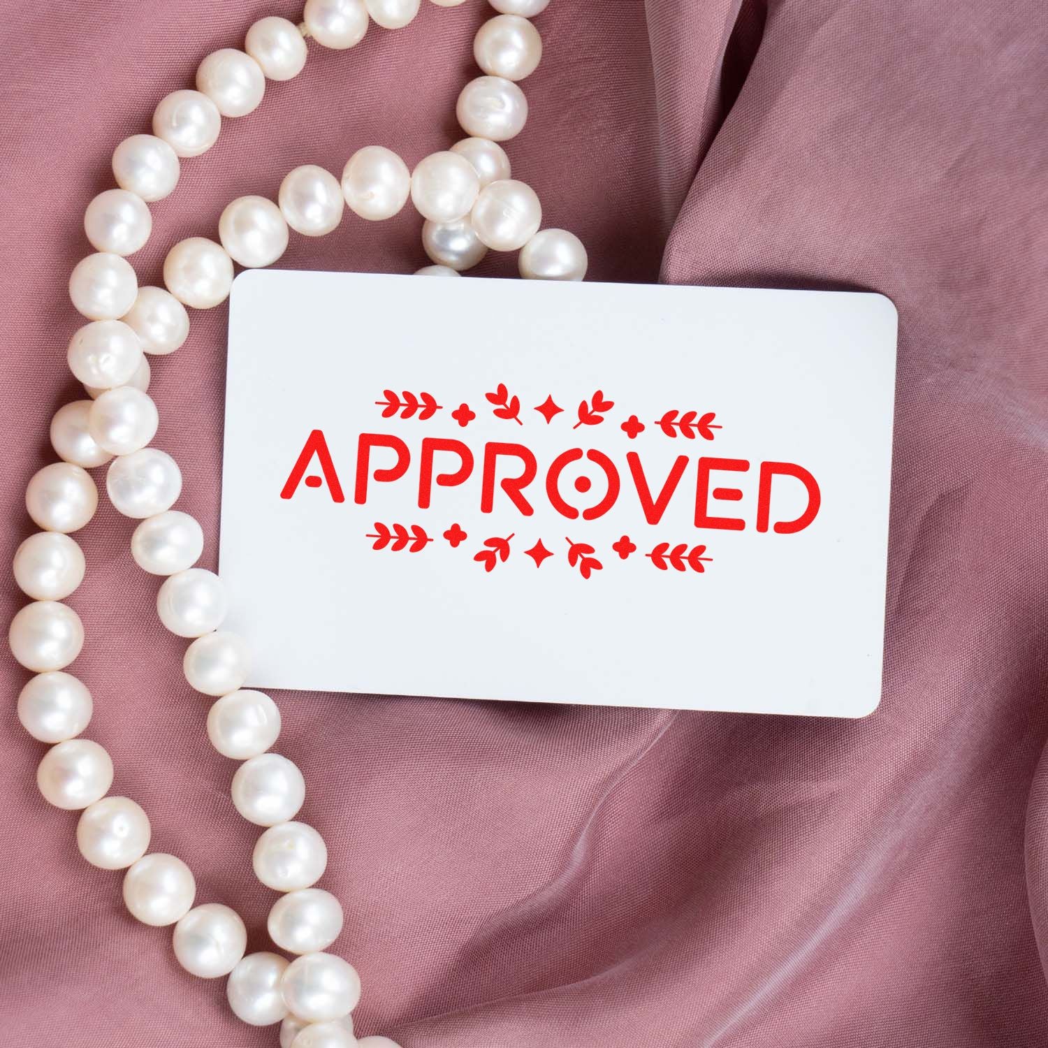 White approval stamp card with red APPROVED seal, pearl strands, and mauve fabric.