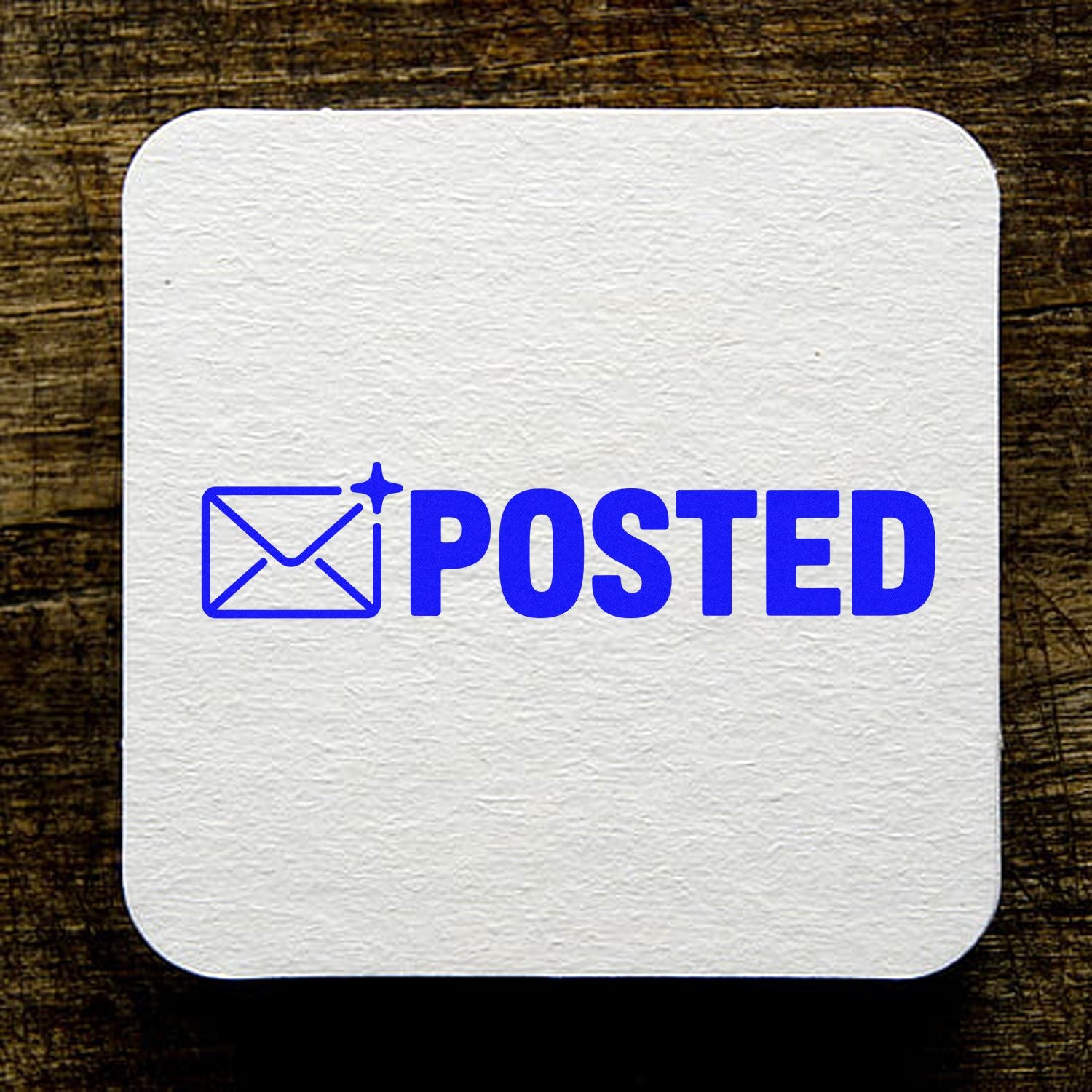 Blue POSTED envelope seal stamp on a white textured square card over dark wood background