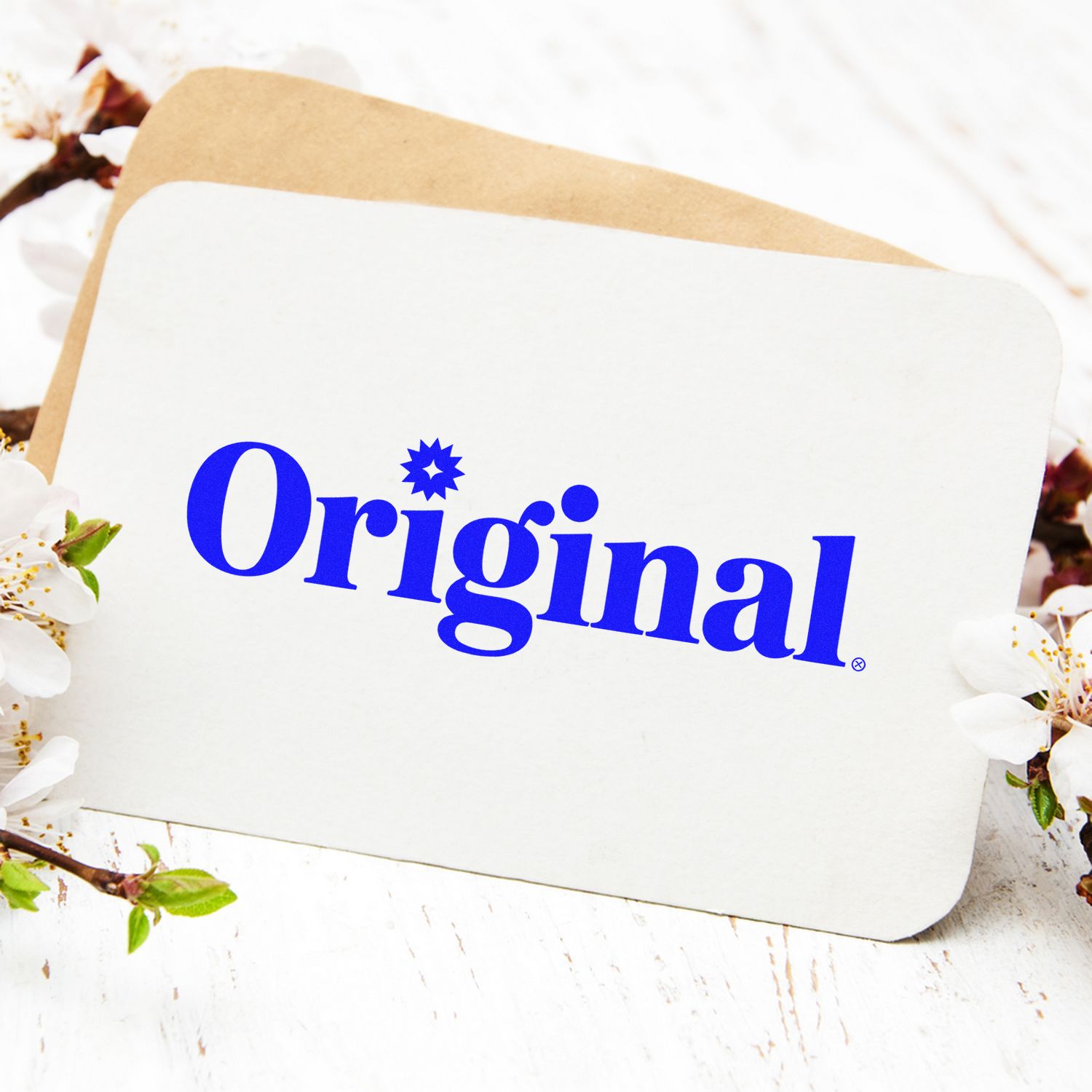 White stamp and seal card with bold blue Original text, beige backing, and white blossoms on a light marble background.
