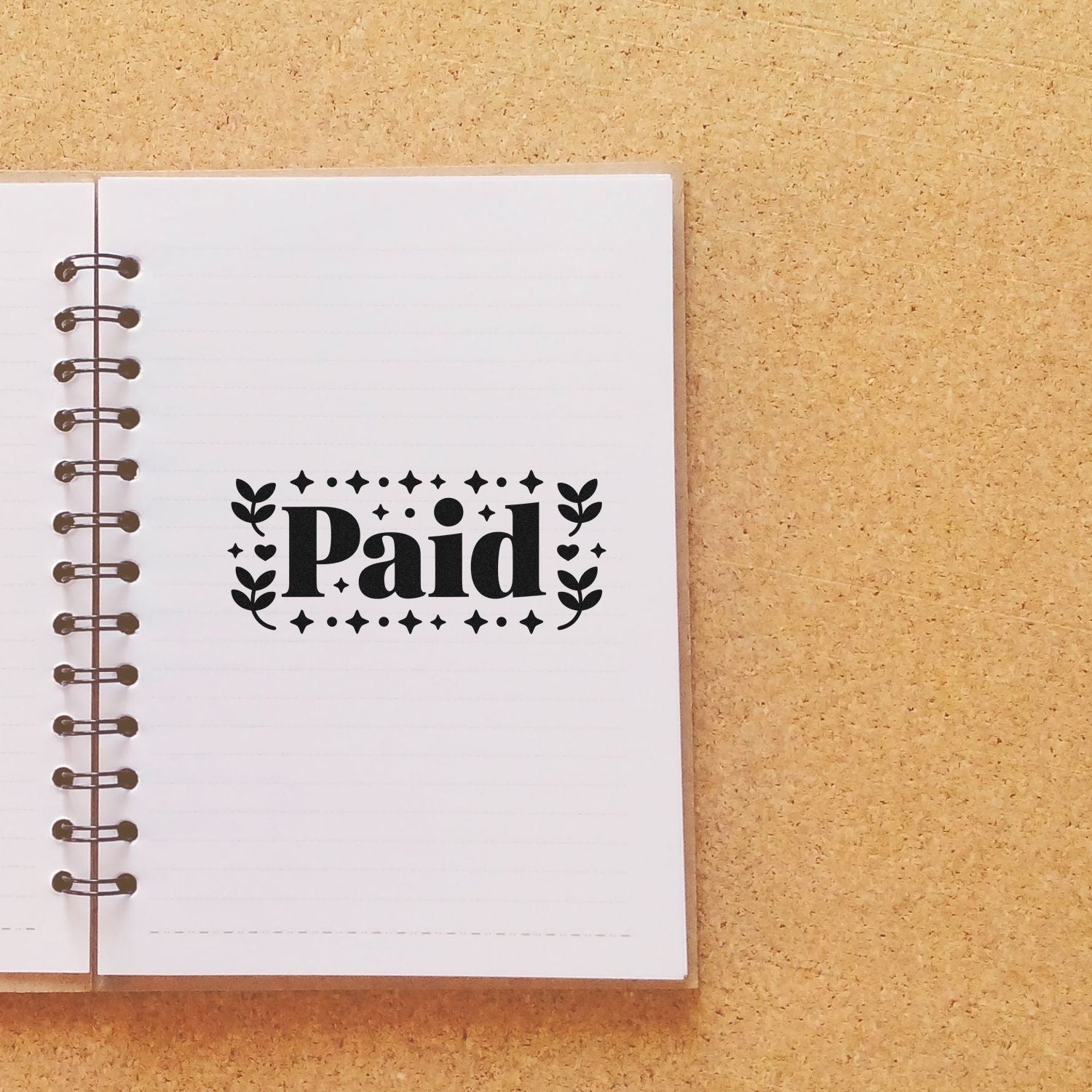 Black Paid stamp seal on a white spiral notebook page, with decorative stars and leaves on a tan cork background.