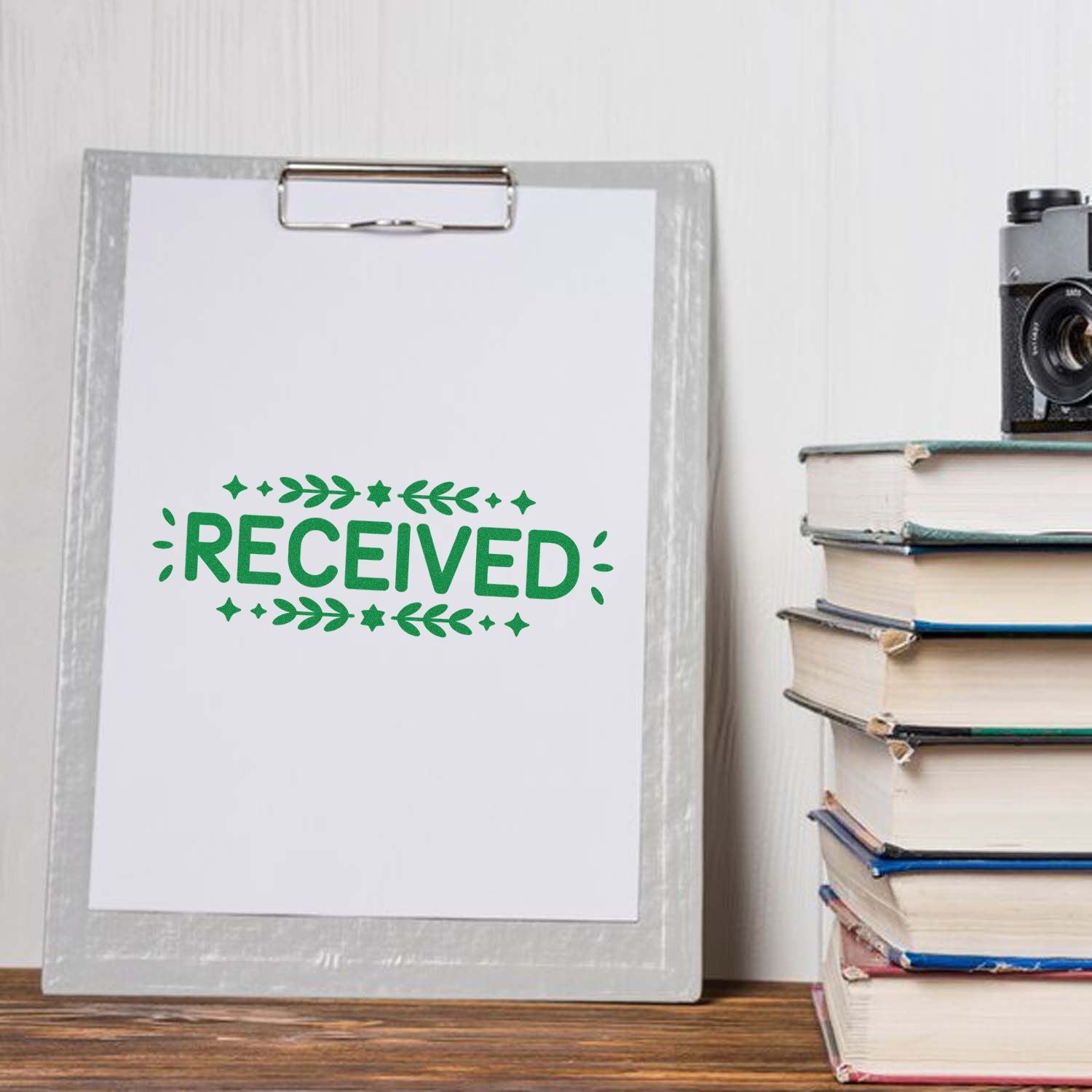 Green RECEIVED rubber stamp and seal on a clipboard beside stacked books and a vintage camera