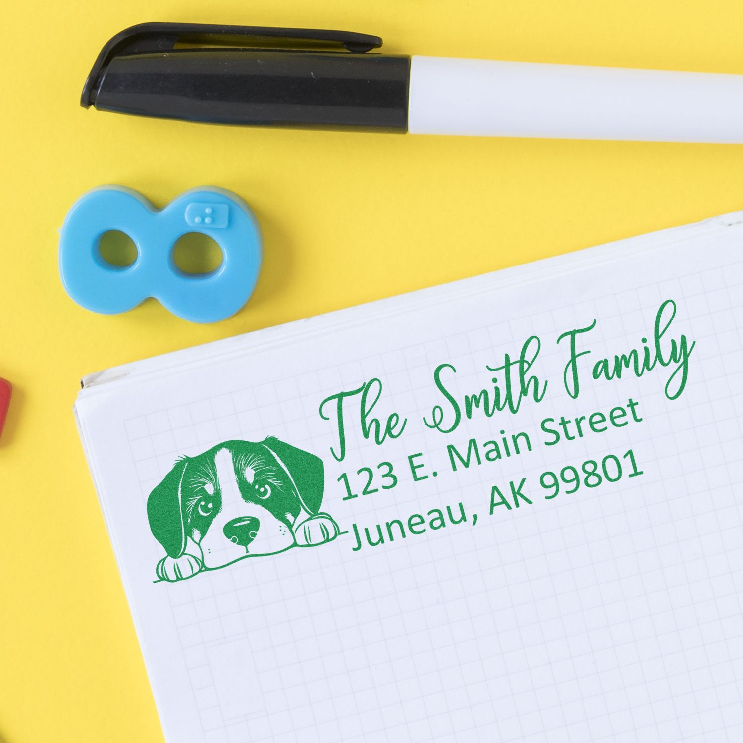 Blue self-inking seal and black marker beside notebook with green custom family address stamp and dog graphic on yellow background