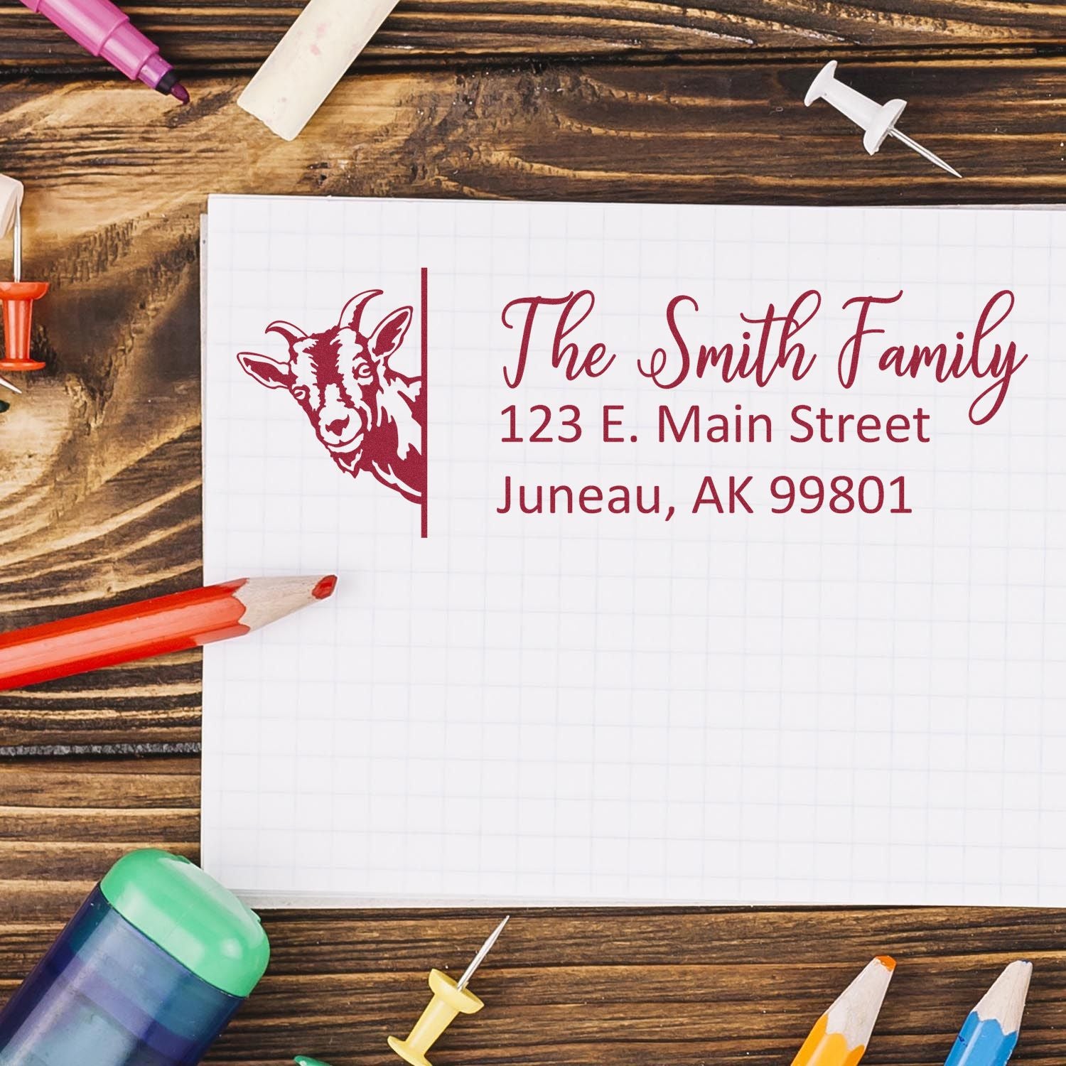 Custom red address stamp and seal with goat logo, The Smith Family, on graph paper with colored pencils on a wood desk.