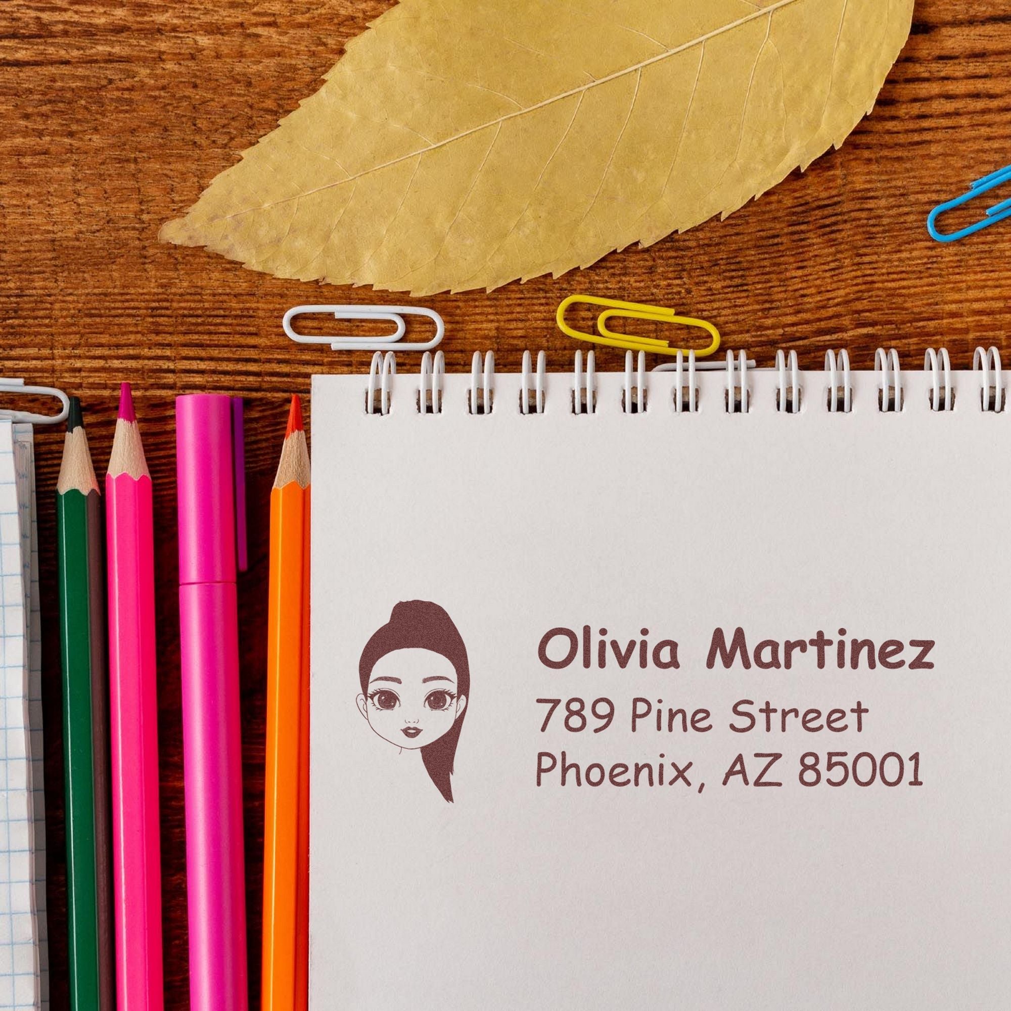 Custom stamp and seal products on a white notebook, with brown address text, girl icon, and colorful pencils on wood.