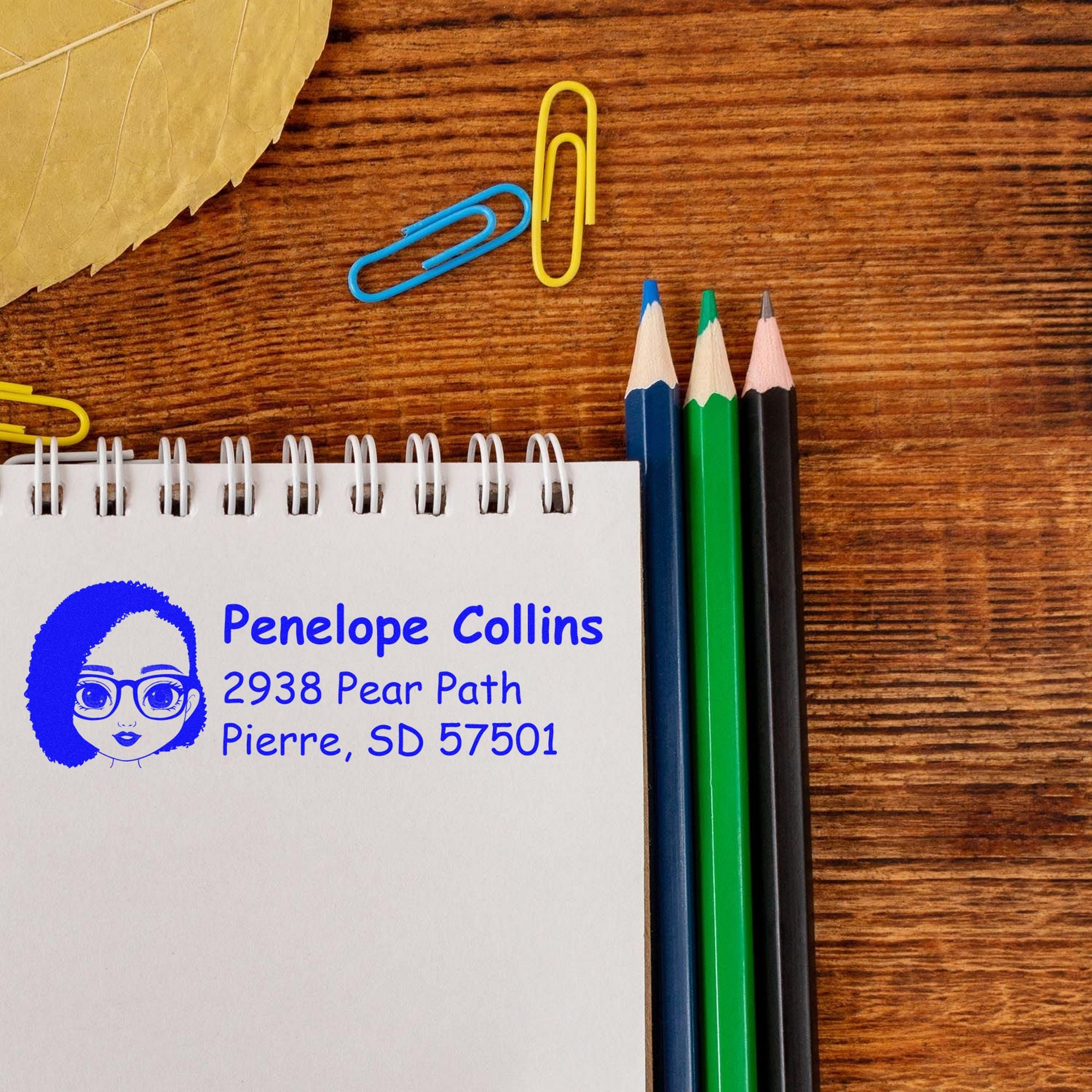 Blue personalized address stamp on a white spiral notebook, with colored pencils and paper clips on a wood desk.