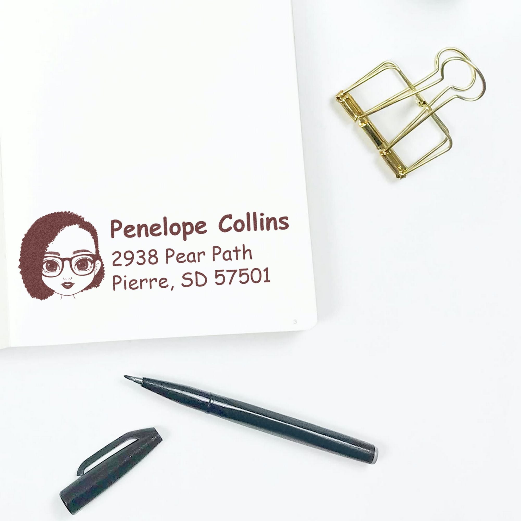 Custom stamp and seal set with burgundy return address stamp, illustrated face, gold binder clip, and black pen on white.