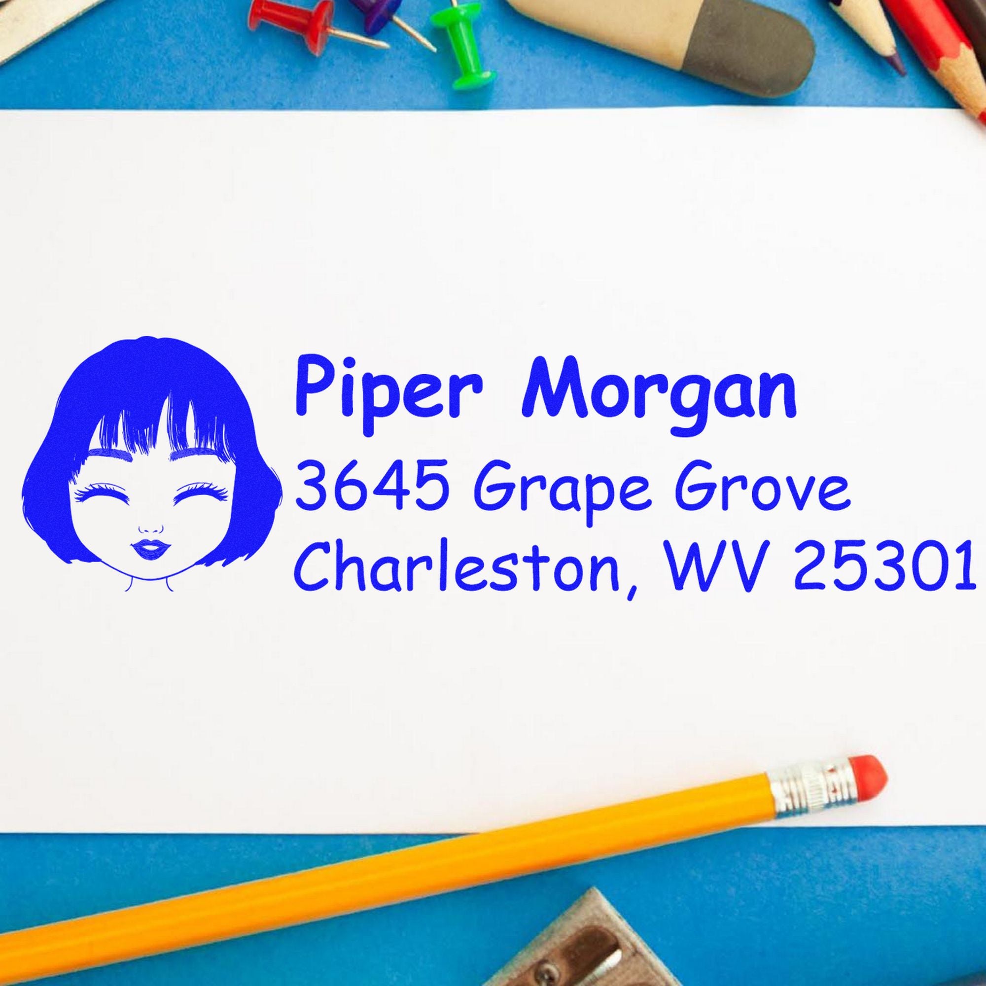 Blue custom address stamp with woman face icon and Piper Morgan address, on white paper with pencils and push pins.