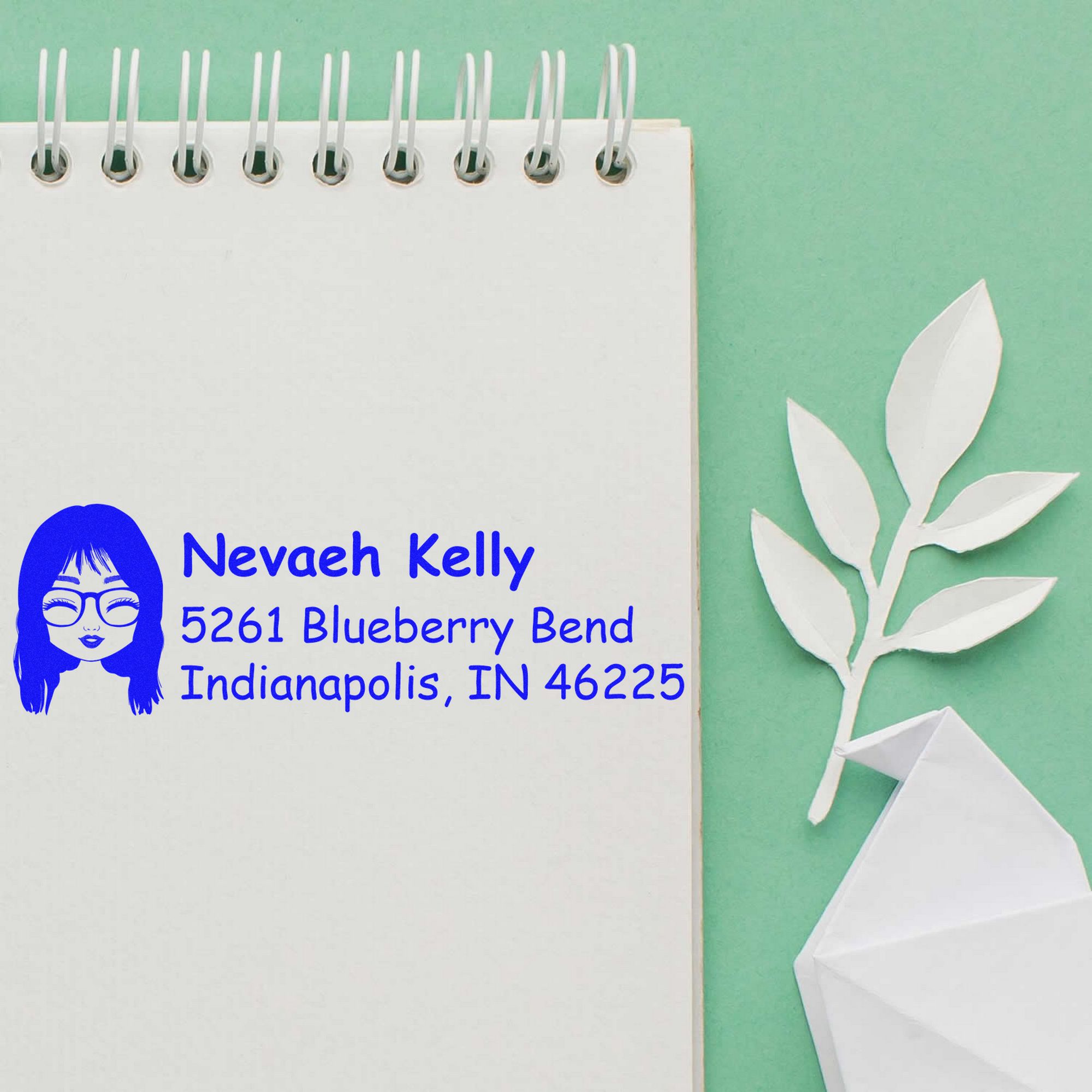 Custom blue address stamp on white spiral notebook, with white paper leaf seal accents on mint green background.