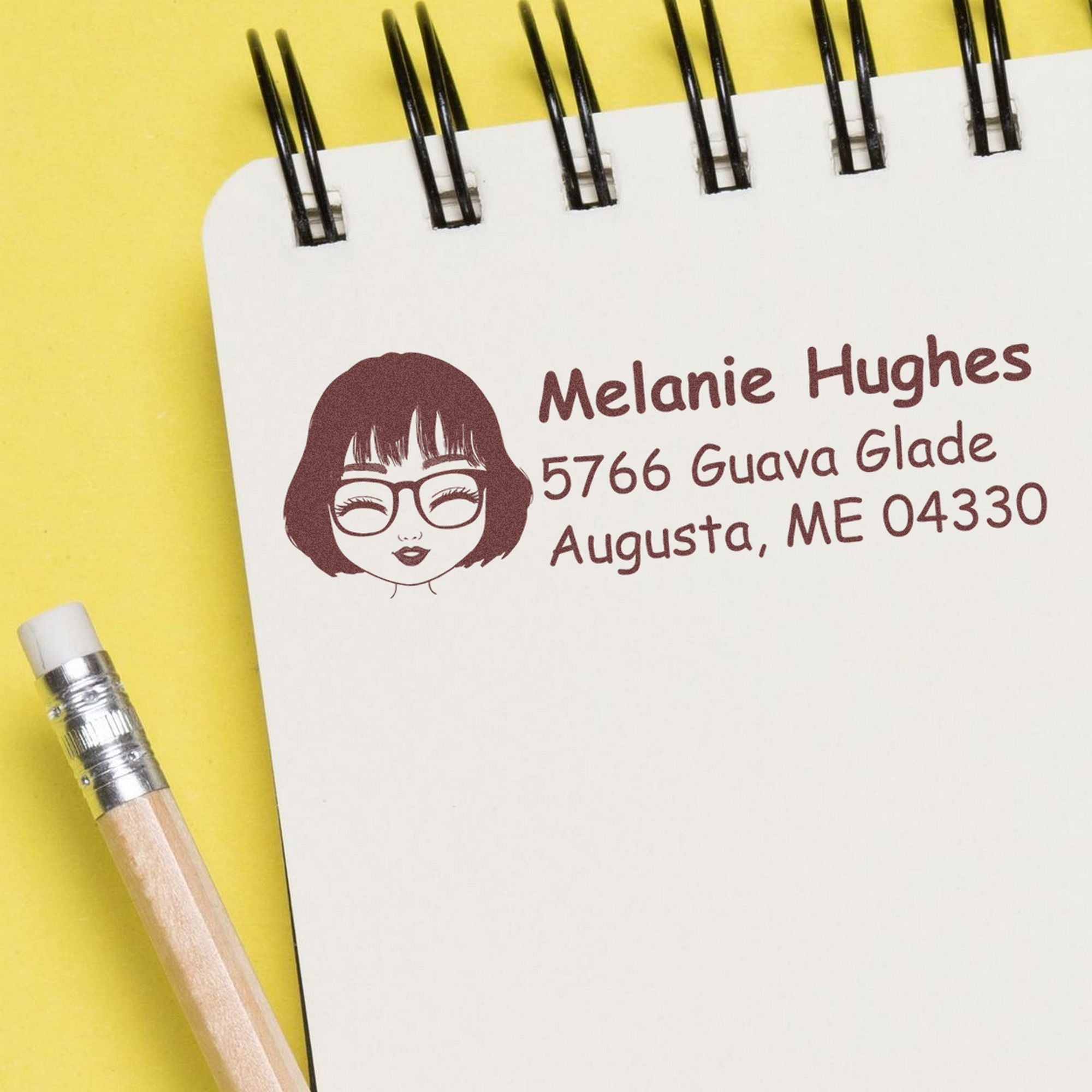 Maroon custom address stamp and seal on a white spiral notebook, with a yellow background and pencil.