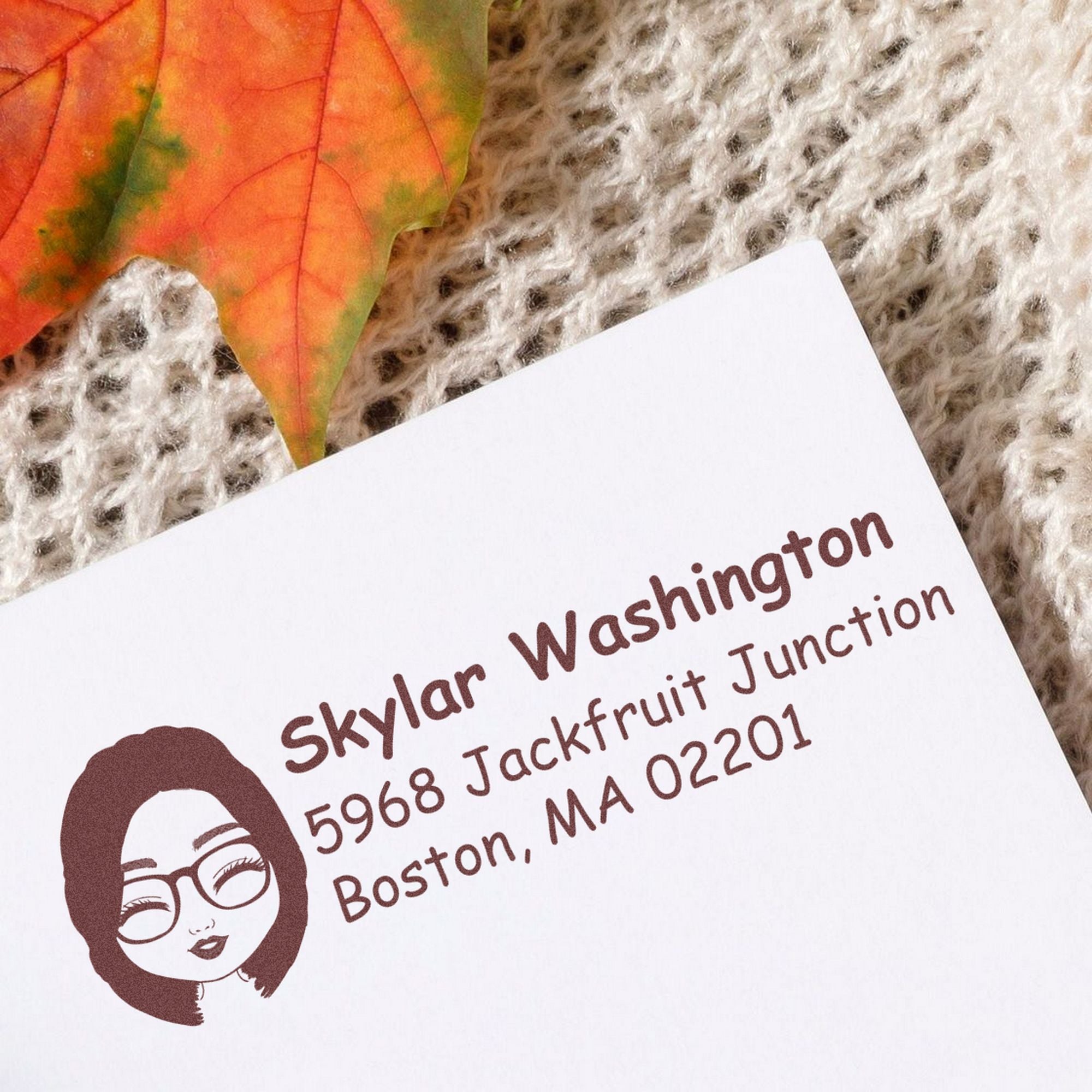 Brown custom stamp and seal on white card, with logo and text, beside an orange autumn leaf on textured knit fabric.