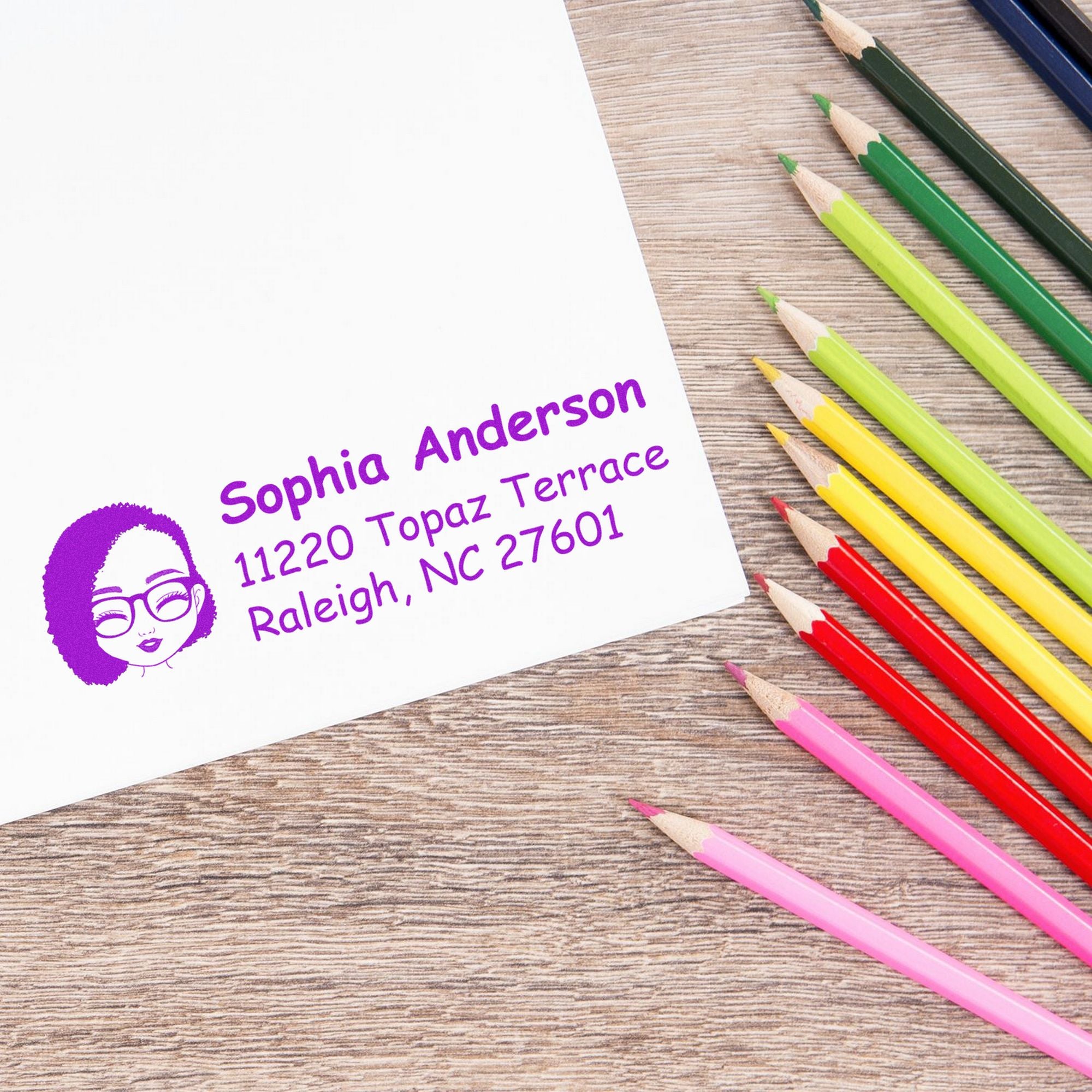 Purple custom address stamp and seal on white paper, beside colorful pencils on a wood desk