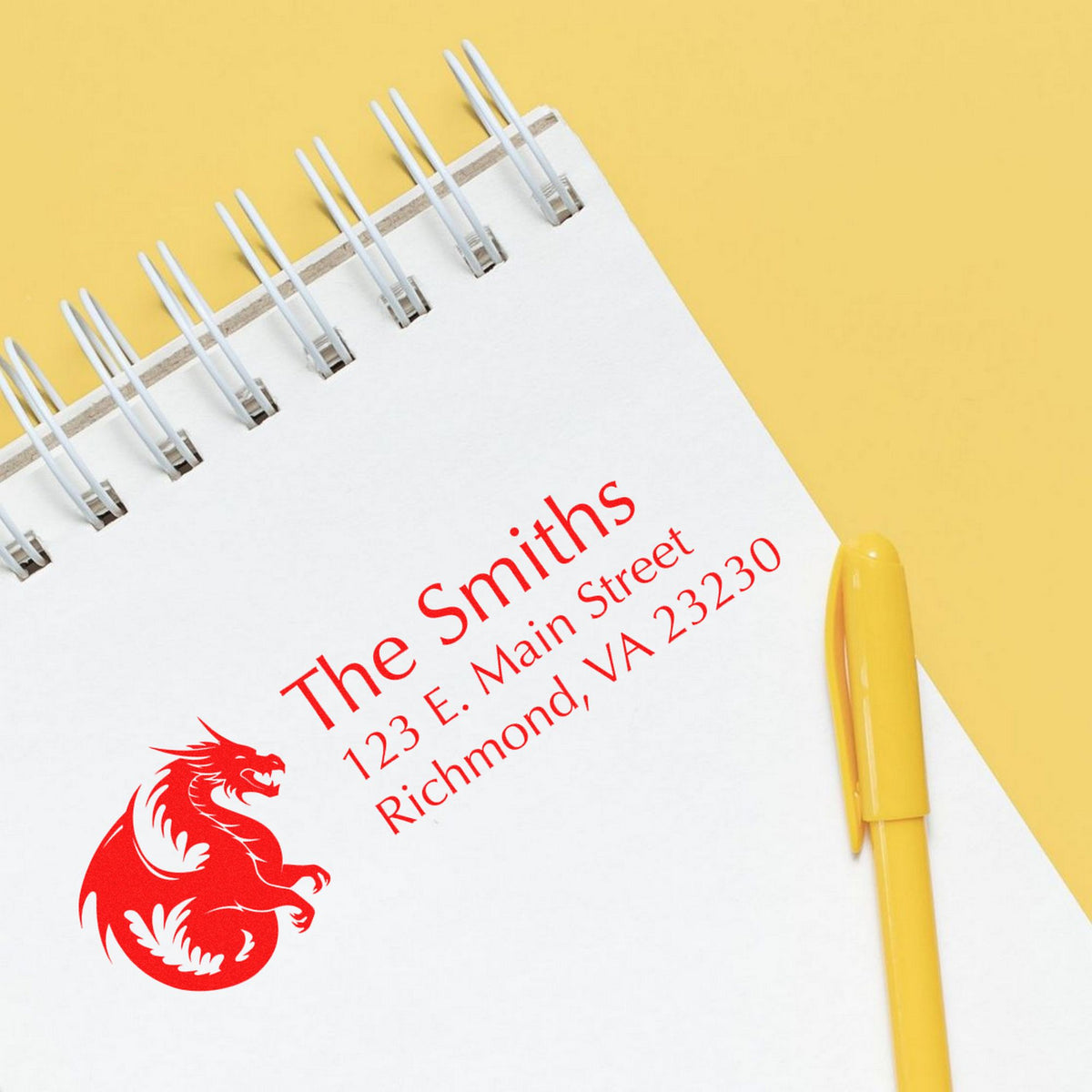 White spiral notebook showing a red custom stamp and seal design with address text, on a yellow background beside a yellow pen.