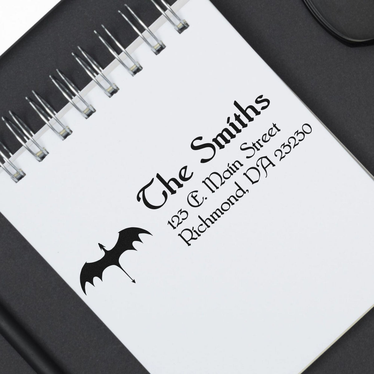 White spiral-bound stamp and seal sample with black custom text and bat logo on a dark background.
