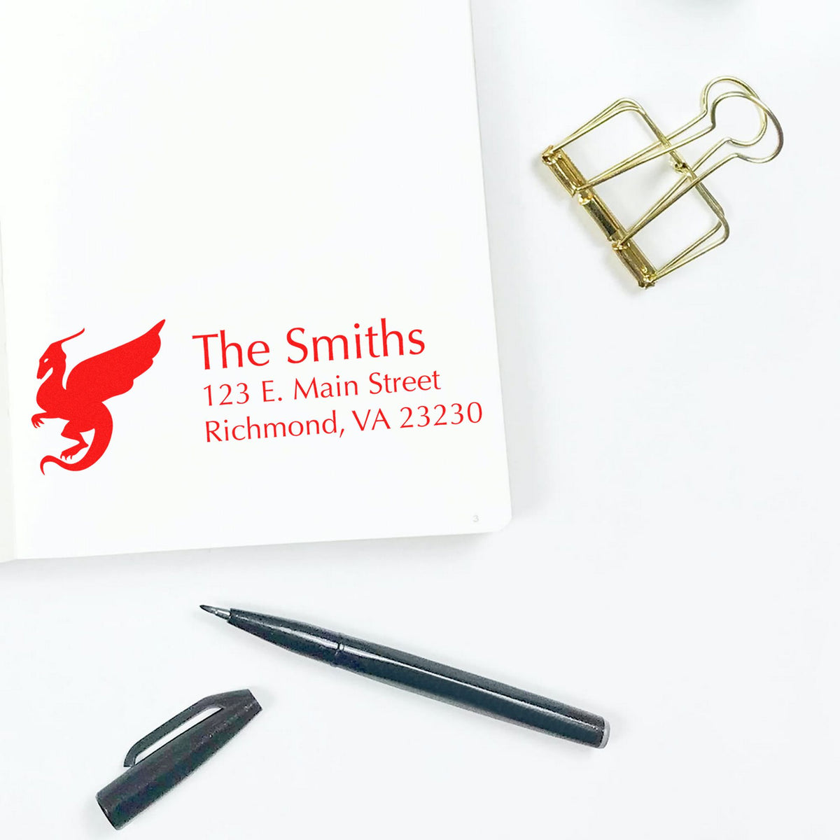 Red custom address stamp impression with griffin logo on white paper, beside gold binder clip and black pen nib.