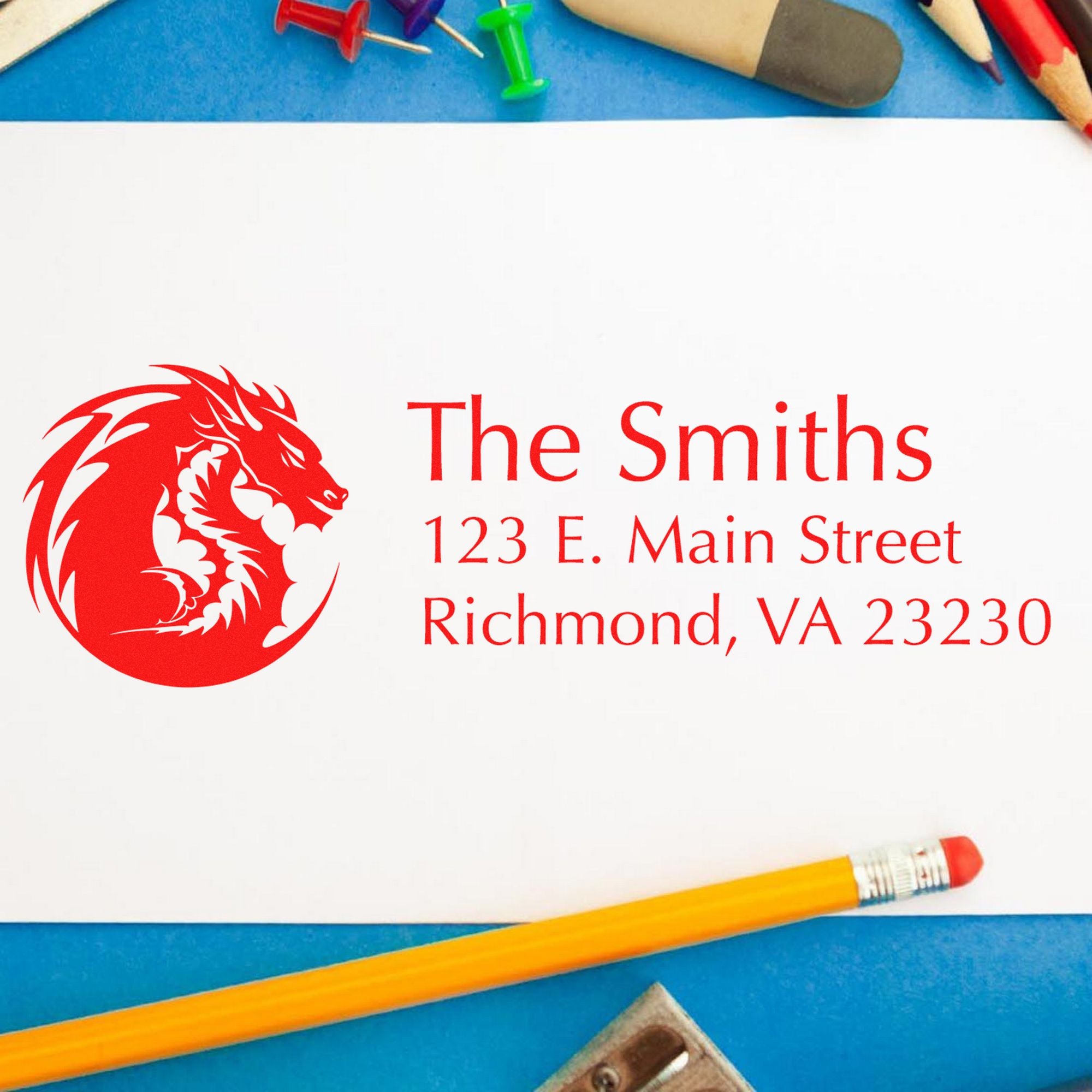 Red custom address stamp and seal with dragon logo, The Smiths address, on white paper with pencils and blue background.