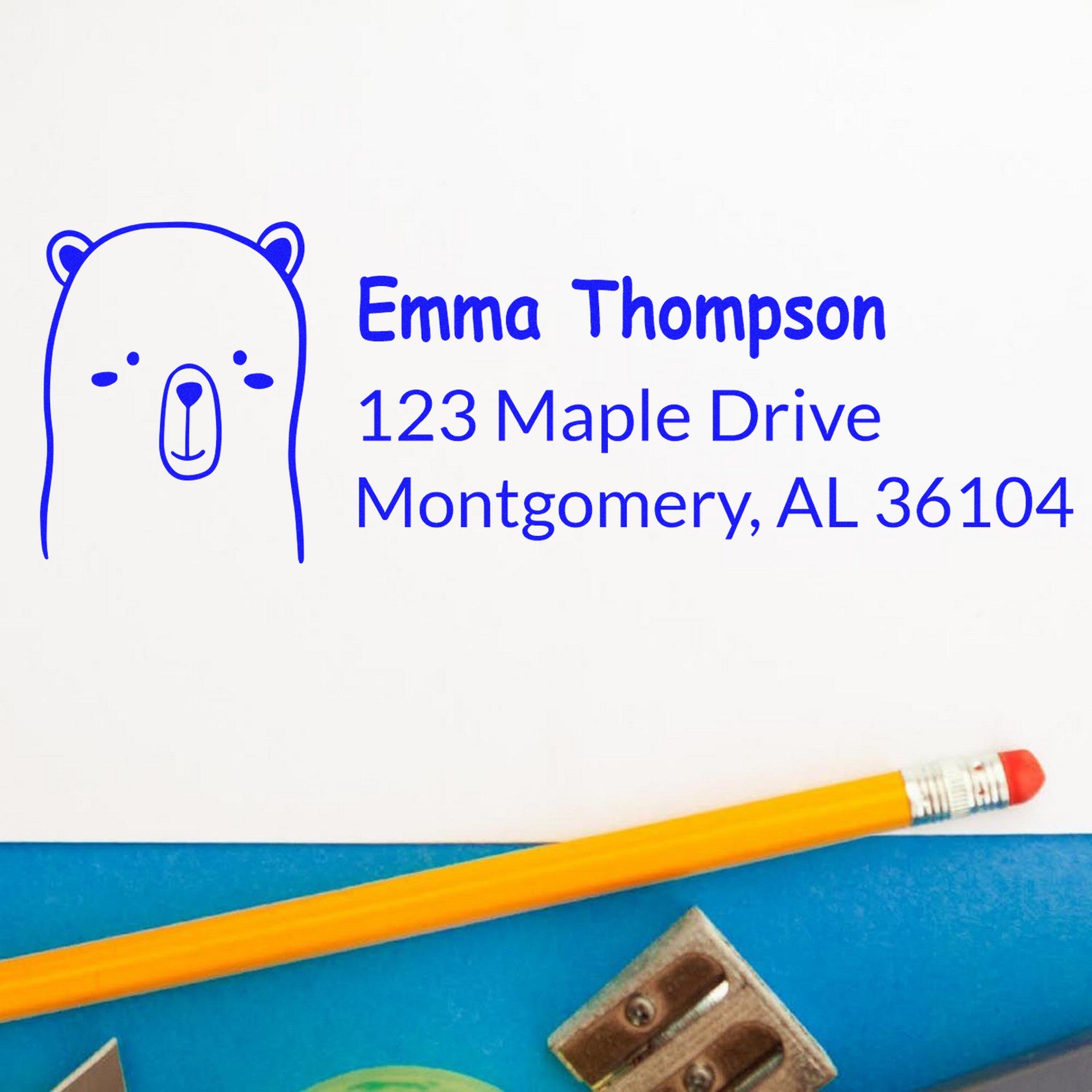 Image of a customizable new address stamper featuring a wood handle and an adorable bear design. The stamp displays the name "Emma Thompson" with the address "123 Maple Drive, Montgomery, AL 36104" in blue ink, next to a pencil and paper clips.