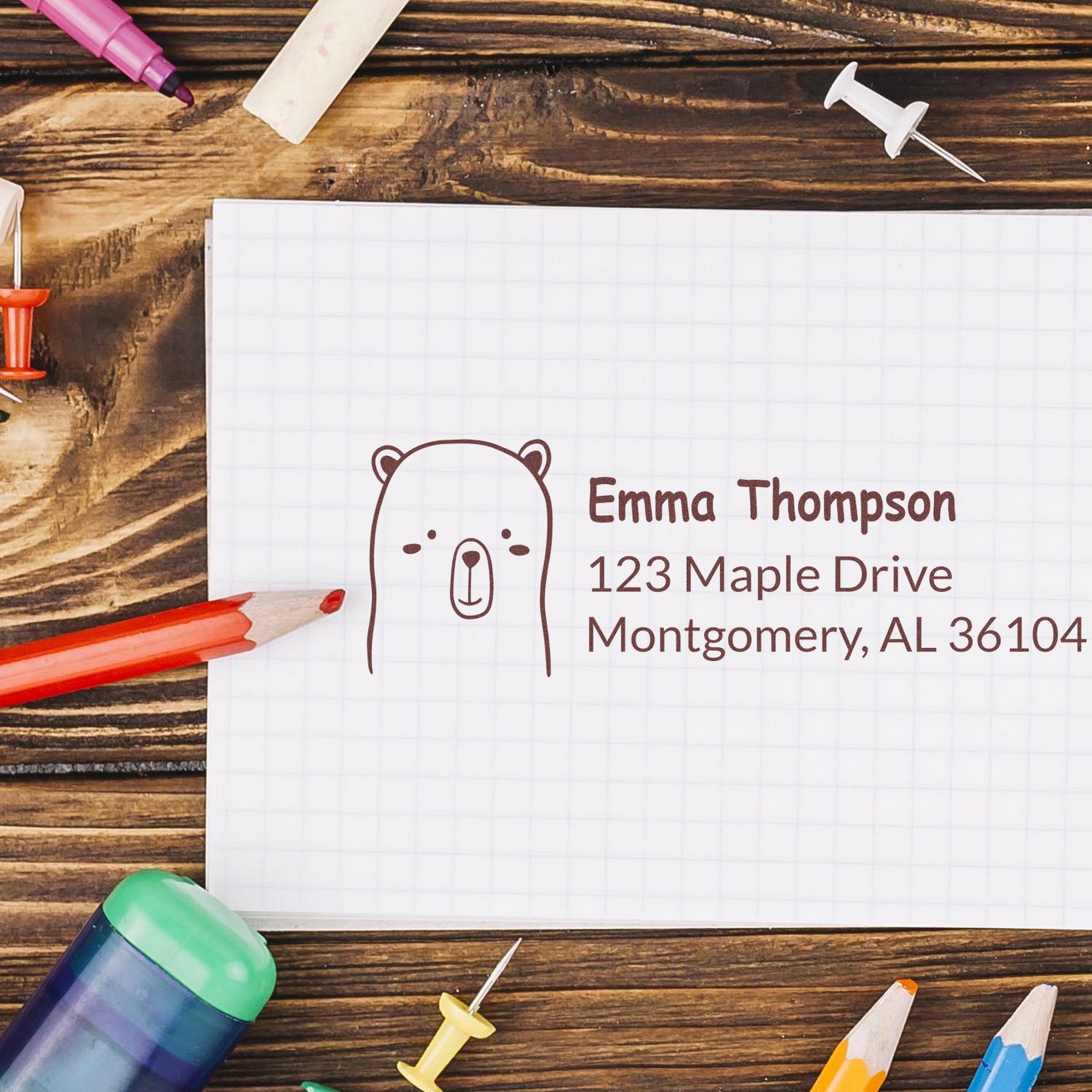 PSI Adorable Bear Customized Address Return Pre-Inked Stamp on a notepad with the name Emma Thompson and address 123 Maple Drive, Montgomery, AL 36104. Surrounded by colorful pencils and push pins, showcasing a Customized Return Mail Stamp design.