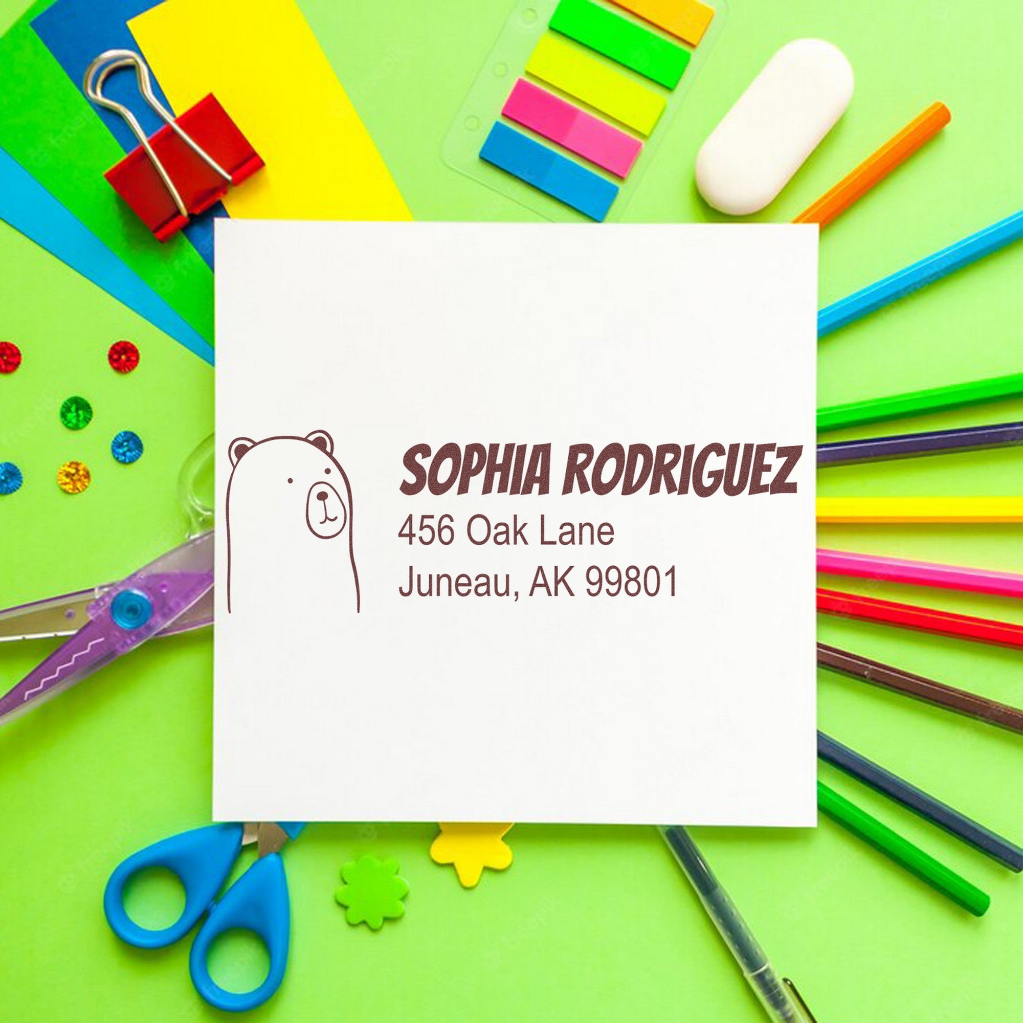 A colorful desk with stationery surrounds a white card featuring a PSI Cuddly Polar Bear Customized Mailing Address Pre-Inked Stamp. The card displays a bear illustration and text: "Sophia Rodriguez, 456 Oak Lane, Juneau, AK 99801." Ideal for a Customized New Address Stamp.