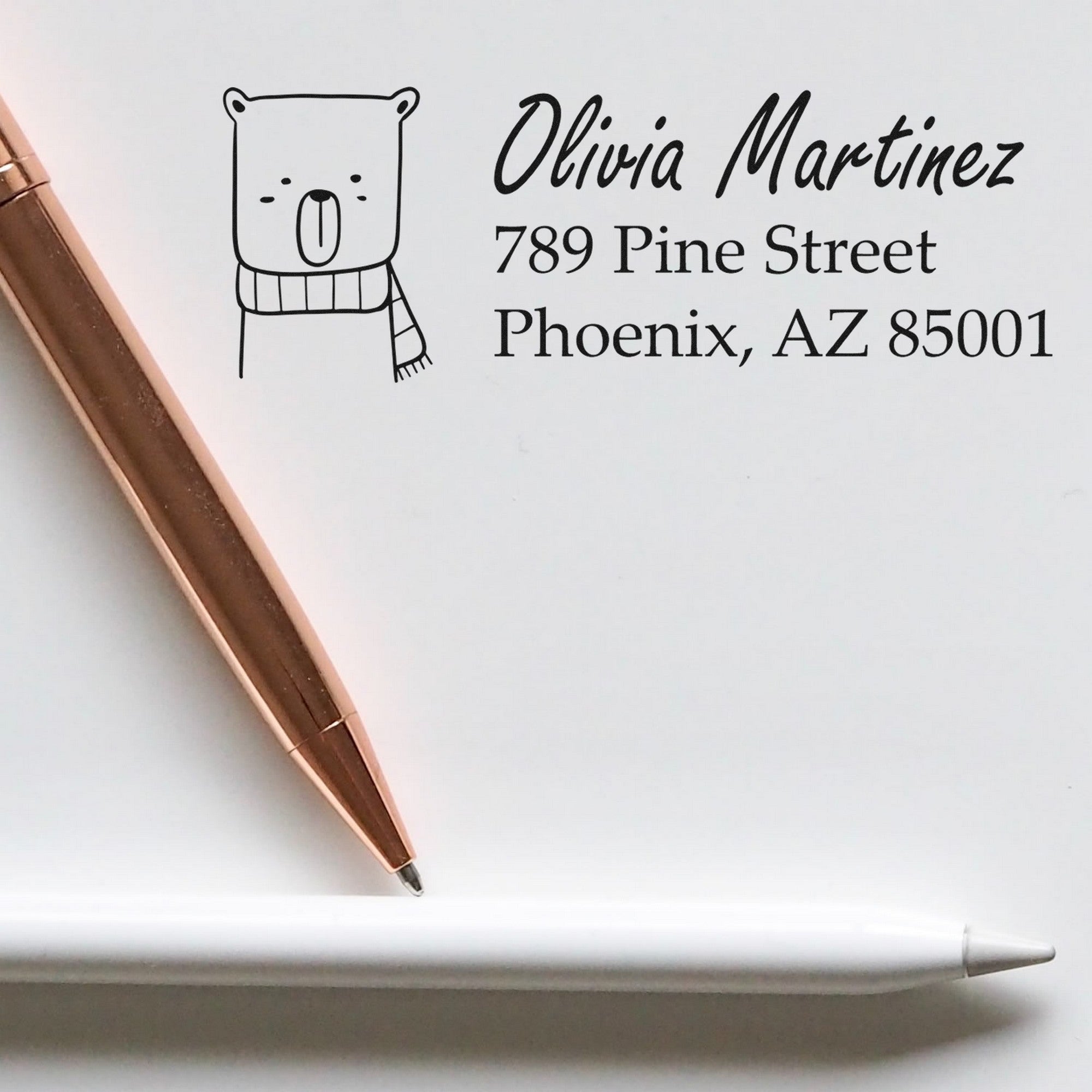 A PSI Snuggly Bear Customized Mail Address Pre-Inked Stamp is shown on an envelope with the name Olivia Martinez, 789 Pine Street, Phoenix, AZ 85001. A rose gold pen and a white pen are placed nearby, highlighting the Personalized Home Stamper design.