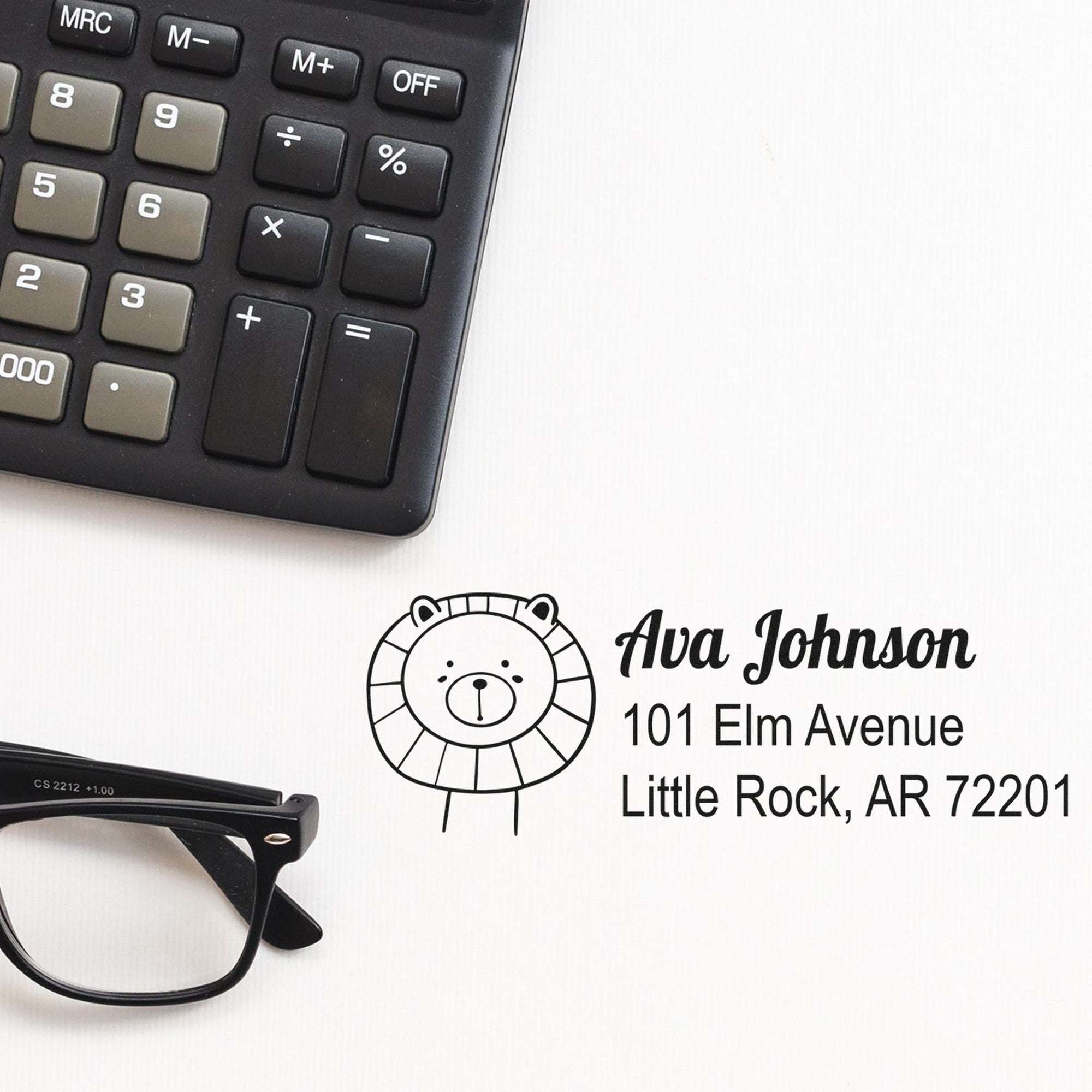 A desk scene featuring a calculator, glasses, and a Lovable Lion's Lair Customize Address Return Self-Inking Stamp. The Customized Mailing Stamper displays "Ava Johnson, 101 Elm Avenue, Little Rock, AR 72201" with a cute lion illustration.