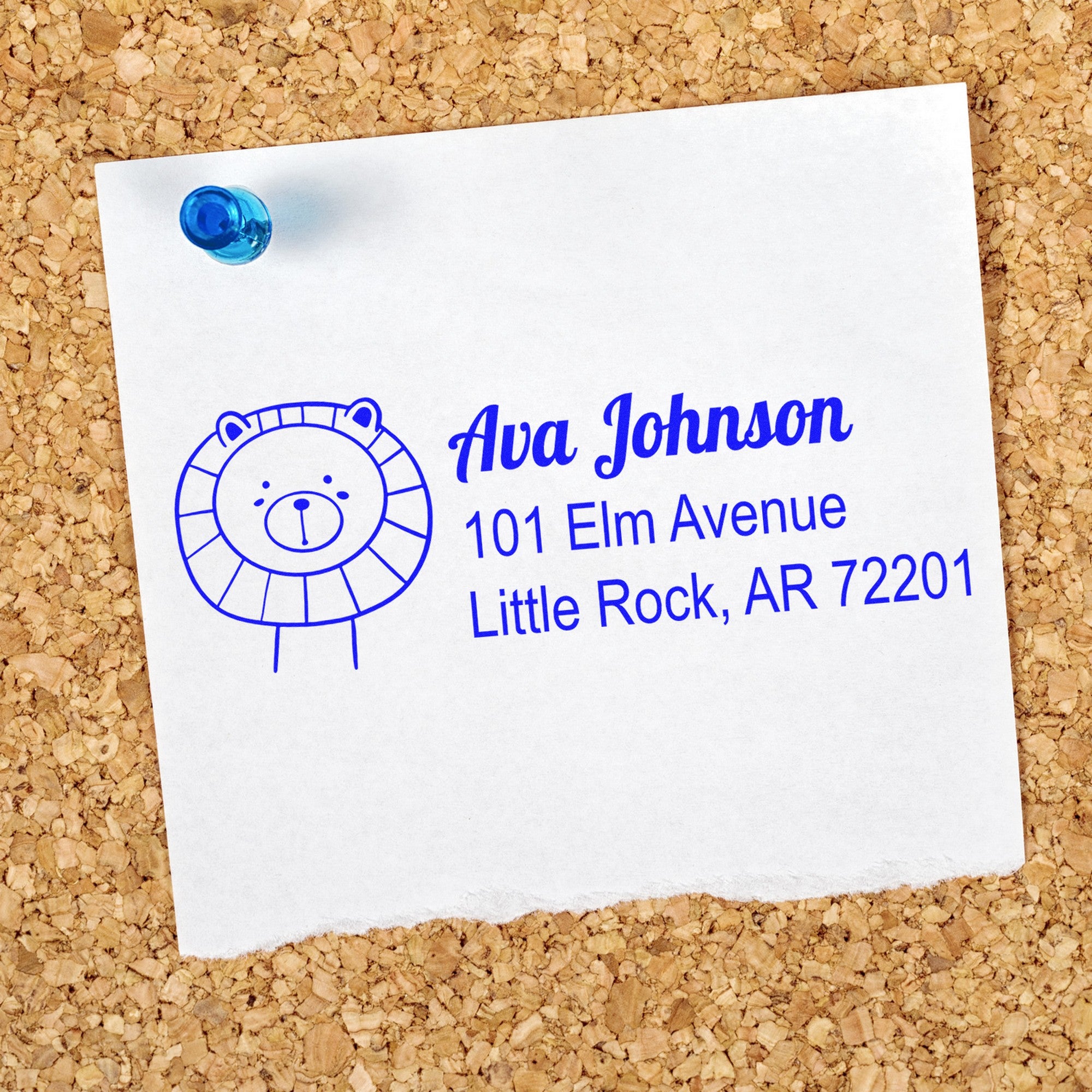 Slim Pre-Inked Lovable Lion's Lair Custom Address Stamp on a corkboard. The personalizable address stamp features a cute lion illustration and text: "Ava Johnson, 101 Elm Avenue, Little Rock, AR 72201" in blue ink on white paper, pinned with a blue pushpin.