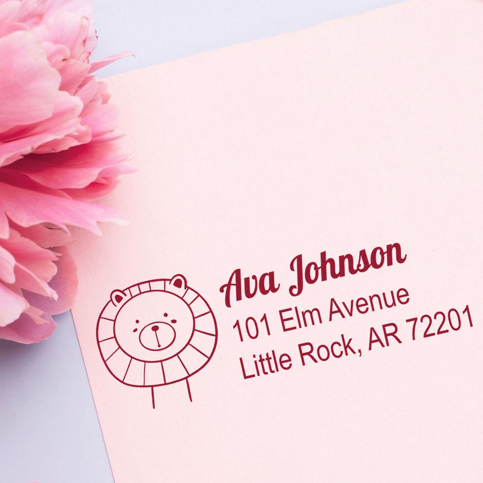 Slim Pre-Inked Lovable Lion's Lair Custom Address Stamp on pink paper with a lion illustration. Text reads "Ava Johnson, 101 Elm Avenue, Little Rock, AR 72201." Pink flower nearby. Customizable Return Mail Stamp for personalized correspondence.