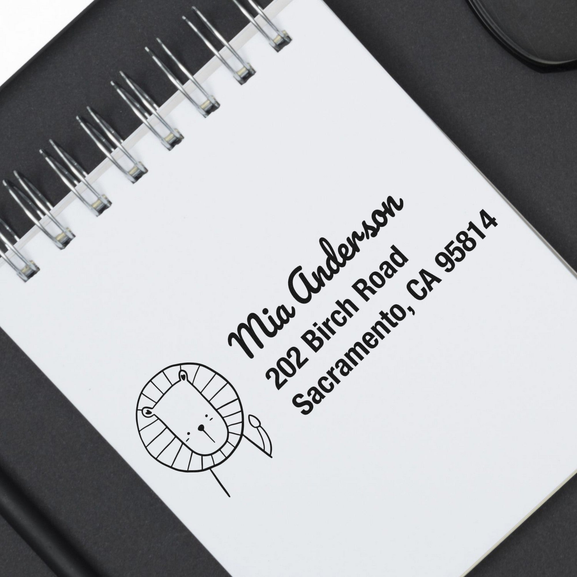 A notebook with a stamped mailing address featuring a lion illustration. The text reads "Mia Anderson, 202 Birch Road, Sacramento, CA 95814." The Little Roar Customize Mailing Address Self-Inking Stamp is used, showcasing a custom-made mailing stamper design.