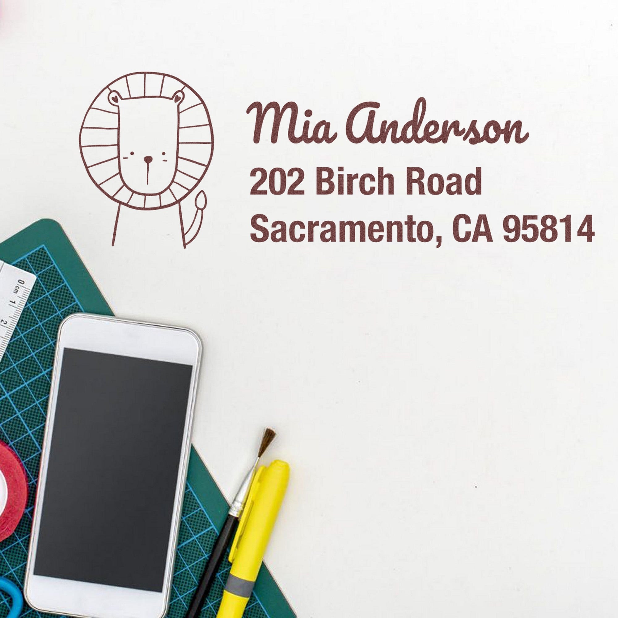 PSI Little Roar Customized Home Address For Envelopes Pre-Inked Stamp featuring a lion design and customizable address stamp with "Mia Anderson, 202 Birch Road, Sacramento, CA 95814" next to stationery items and a smartphone on a desk.