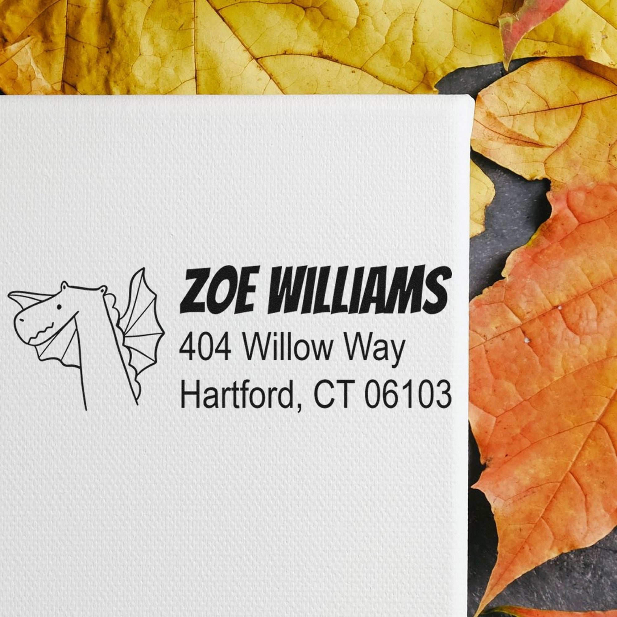 Envelope with Dragon Delight Customize New Address Self-Inking Stamp featuring a dragon design. Text reads 'Zoe Williams, 404 Willow Way, Hartford, CT 06103'. Customizable Mailing Stamp on autumn leaves background.