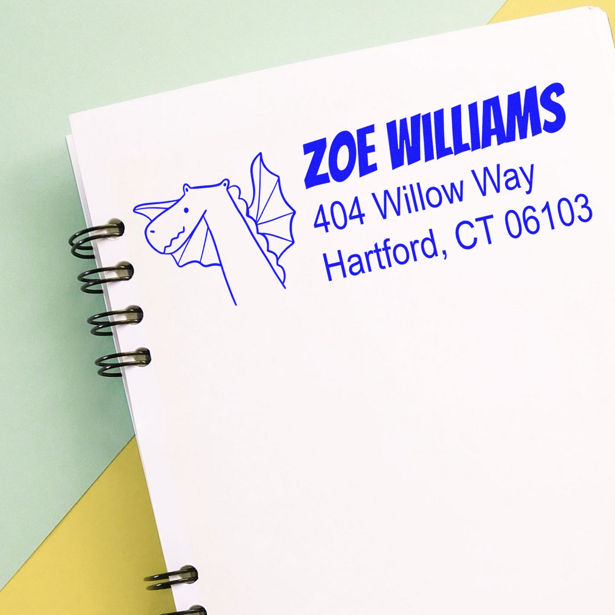 A notebook page featuring a blue Dragon Delight Customize New Address Self-Inking Stamp with a dragon illustration. The Customizable Address Stamper displays "Zoe Williams, 404 Willow Way, Hartford, CT 06103" in bold text.