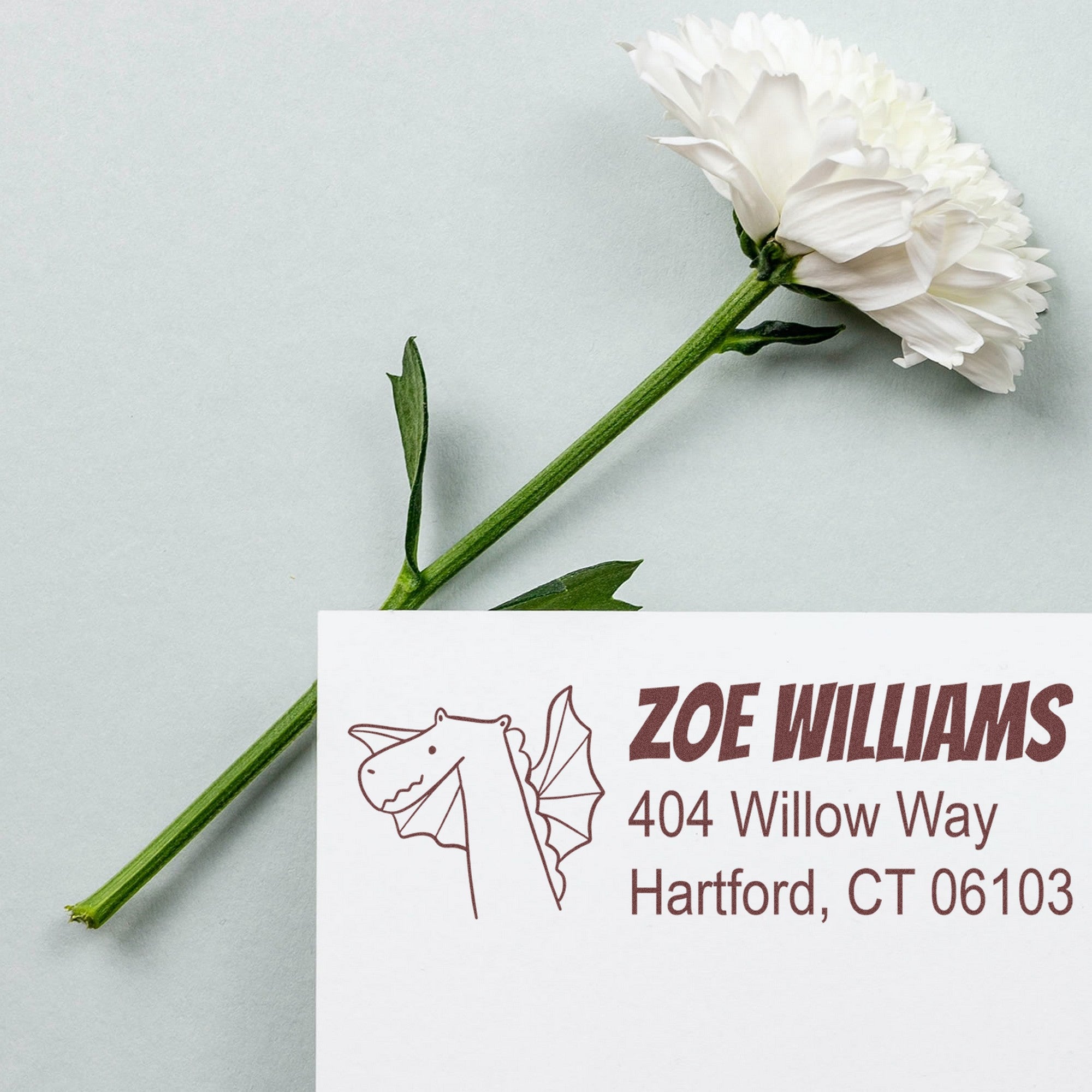 Wood Handle Dragon Delight Personalized Address Return Rubber Stamp on white paper with a dragon illustration and customizable address stamp for Zoe Williams, 404 Willow Way, Hartford, CT 06103. A white flower lies beside the paper on a light background.