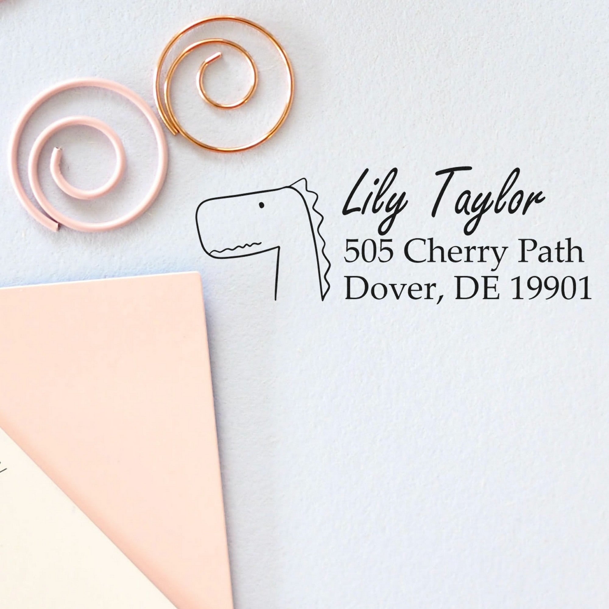 Mirthful Dino Customize Home Address For Envelopes Self-Inking Stamp displayed on a light surface. The stamp features a dinosaur illustration with customizable mailing details, showcasing "Lily Taylor, 505 Cherry Path, Dover, DE 19901" alongside decorative spiral paper clips.