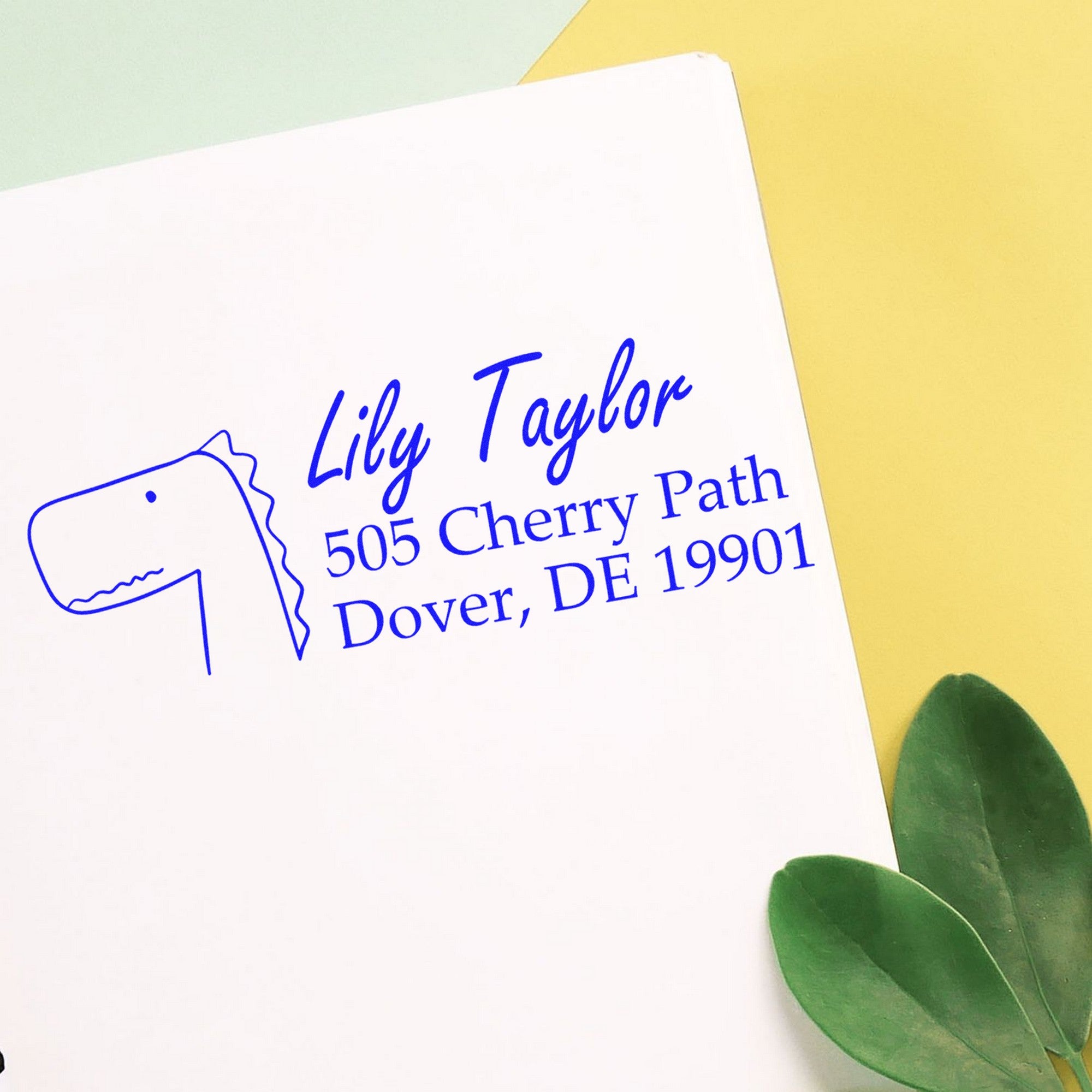 Wood Handle Mirthful Dino Personalized Mailing Address Rubber Stamp on white paper, featuring a dinosaur illustration and text "Lily Taylor, 505 Cherry Path, Dover, DE 19901." Personalizable Return Mail Stamper with green leaves and yellow background.