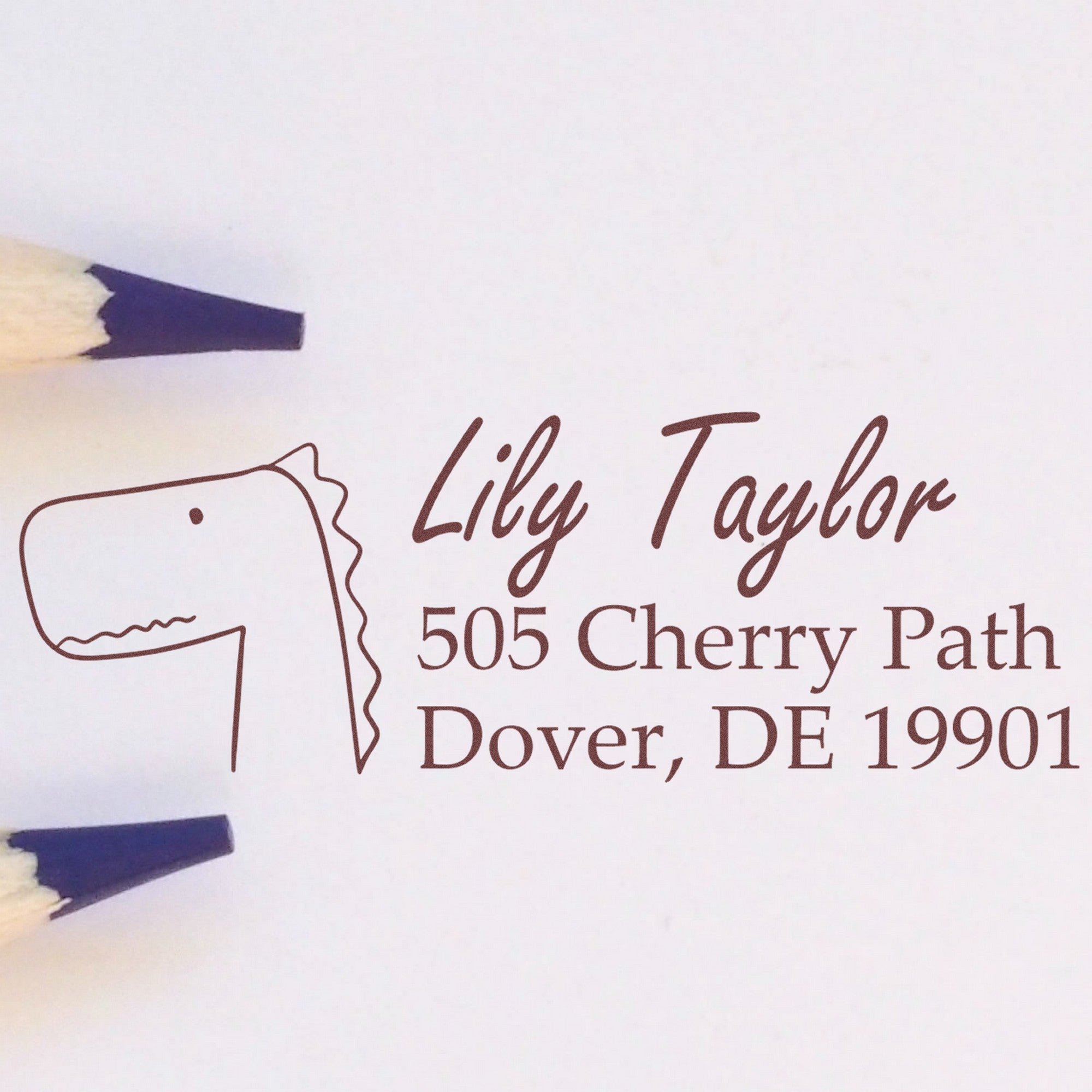 Wood Handle Mirthful Dino Personalized Mailing Address Rubber Stamp with a dinosaur illustration and customizable text. The stamp displays "Lily Taylor, 505 Cherry Path, Dover, DE 19901" and is a personalizable mailing stamp, shown next to two pencils.