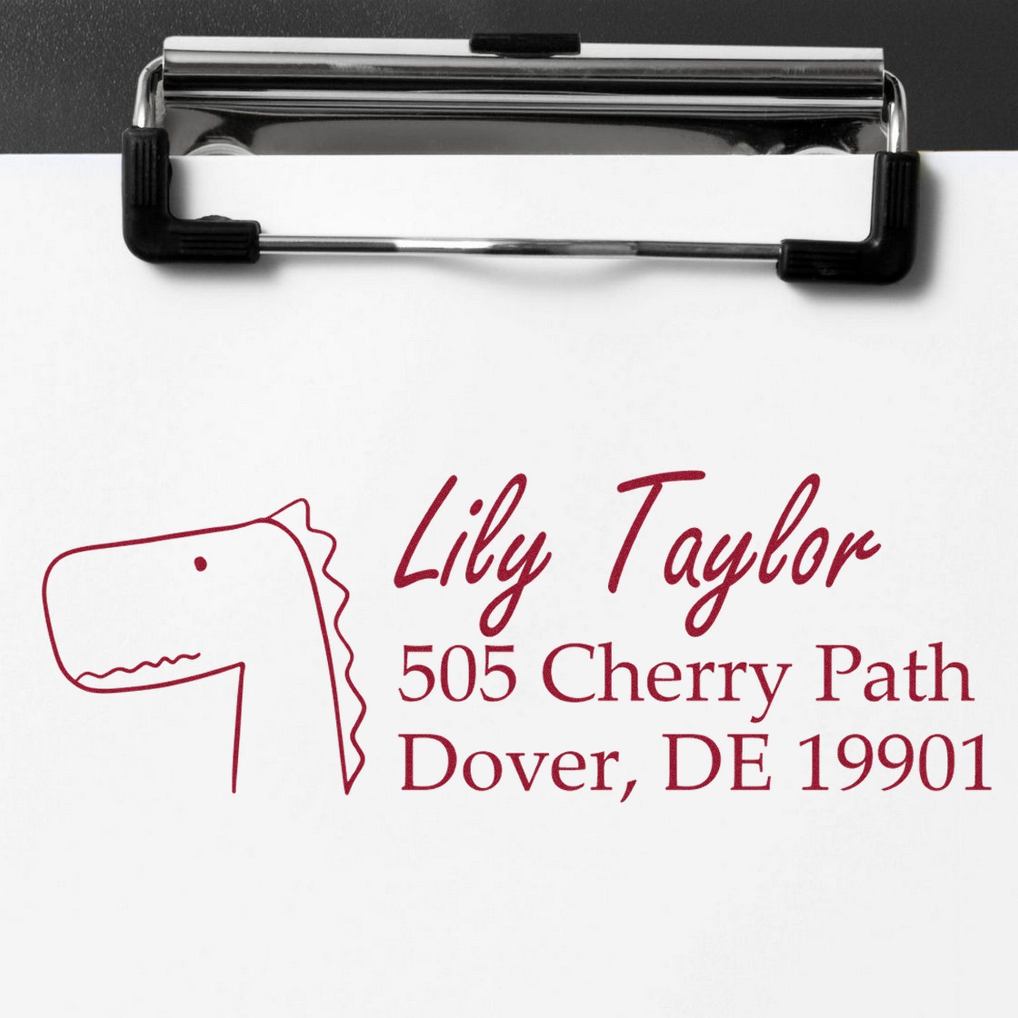 Wood Handle Mirthful Dino Personalized Mailing Address Rubber Stamp on paper, featuring a dinosaur sketch and text: "Lily Taylor, 505 Cherry Path, Dover, DE 19901." This Personalized Address Stamper is shown on a clipboard.