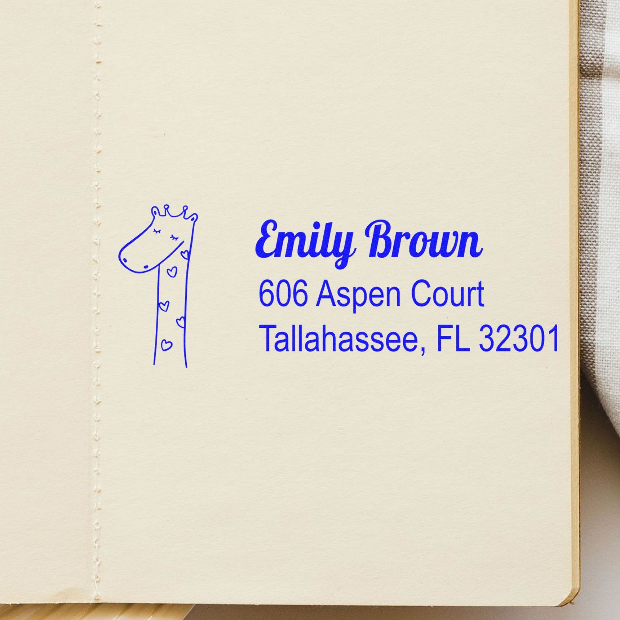 Wood handle lovely giraffe personalized mail address rubber stamp on a notebook page. The custom-made mailing stamper features a cute giraffe design with hearts, alongside the name "Emily Brown" and address "606 Aspen Court, Tallahassee, FL 32301" in blue ink.