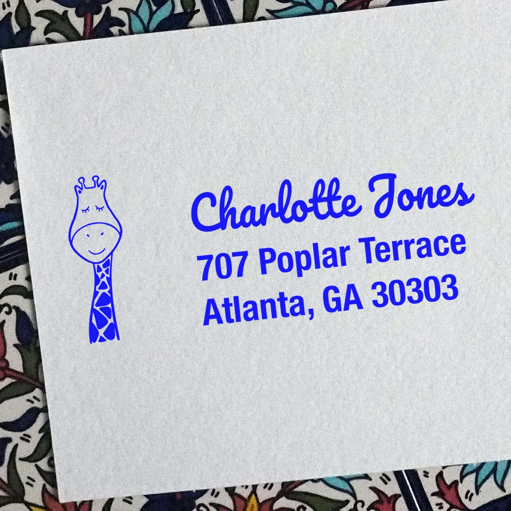 A white envelope features a blue PSI Jolly Giraffe Handmade Mailing Pre-Inked Stamp with a giraffe illustration. The Customized Address Stamp displays "Charlotte Jones, 707 Poplar Terrace, Atlanta, GA 30303" in bold blue text on a patterned background.