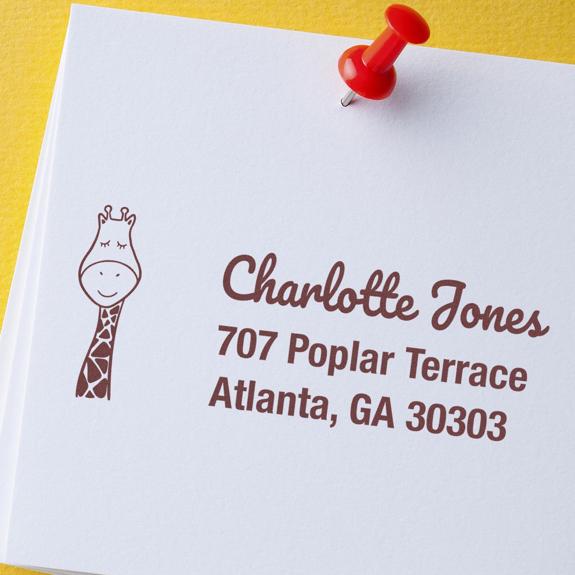 Wood Handle Jolly Giraffe Personalized New Address Rubber Stamp on white paper with a giraffe illustration and text 'Charlotte Jones, 707 Poplar Terrace, Atlanta, GA 30303.' A red pushpin secures the paper. Ideal for a Personalized Home Stamper.