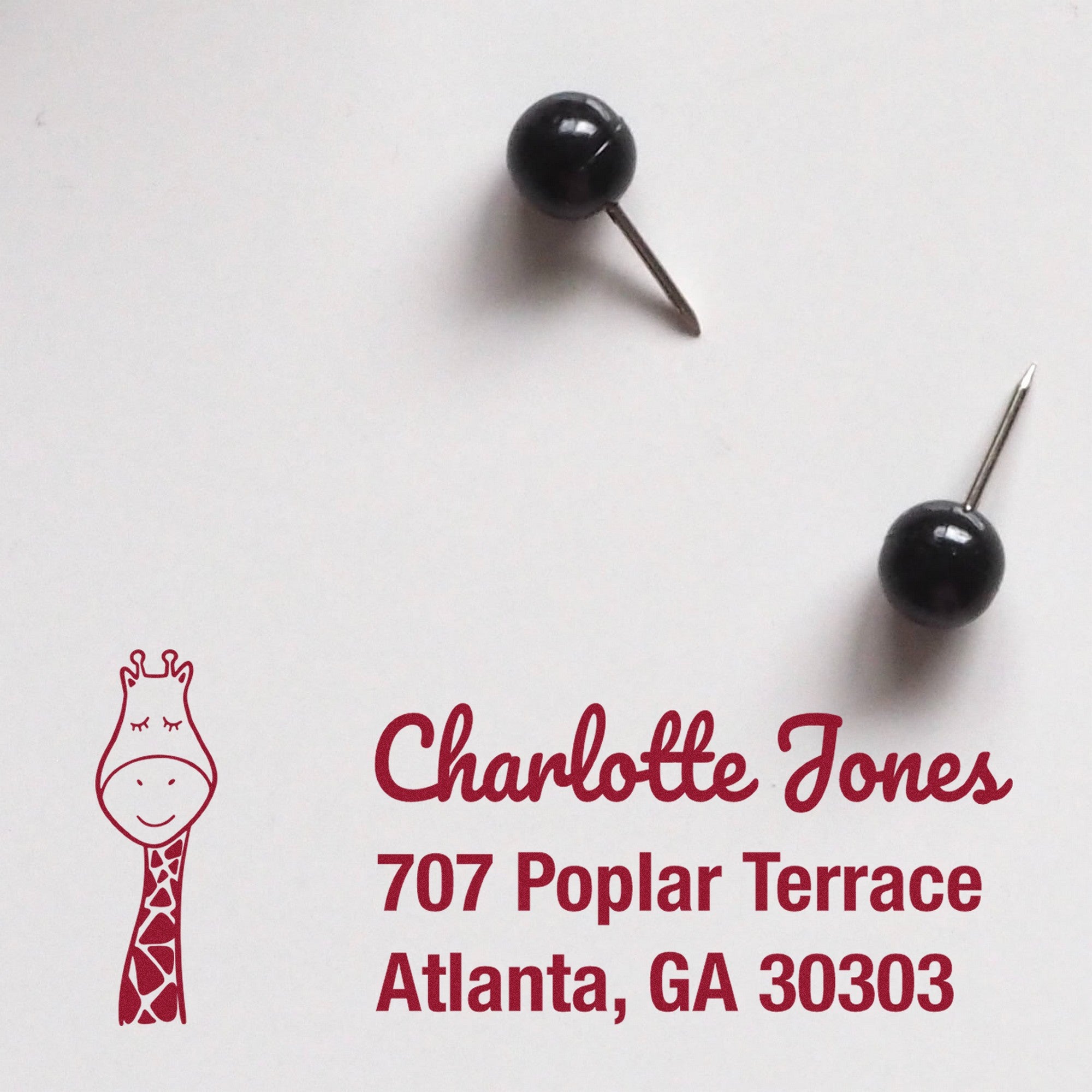 Image of a personalized mailing stamp featuring a wood handle Jolly Giraffe design. The stamp displays the name "Charlotte Jones" and address "707 Poplar Terrace, Atlanta, GA 30303" in red ink, with two black push pins nearby.