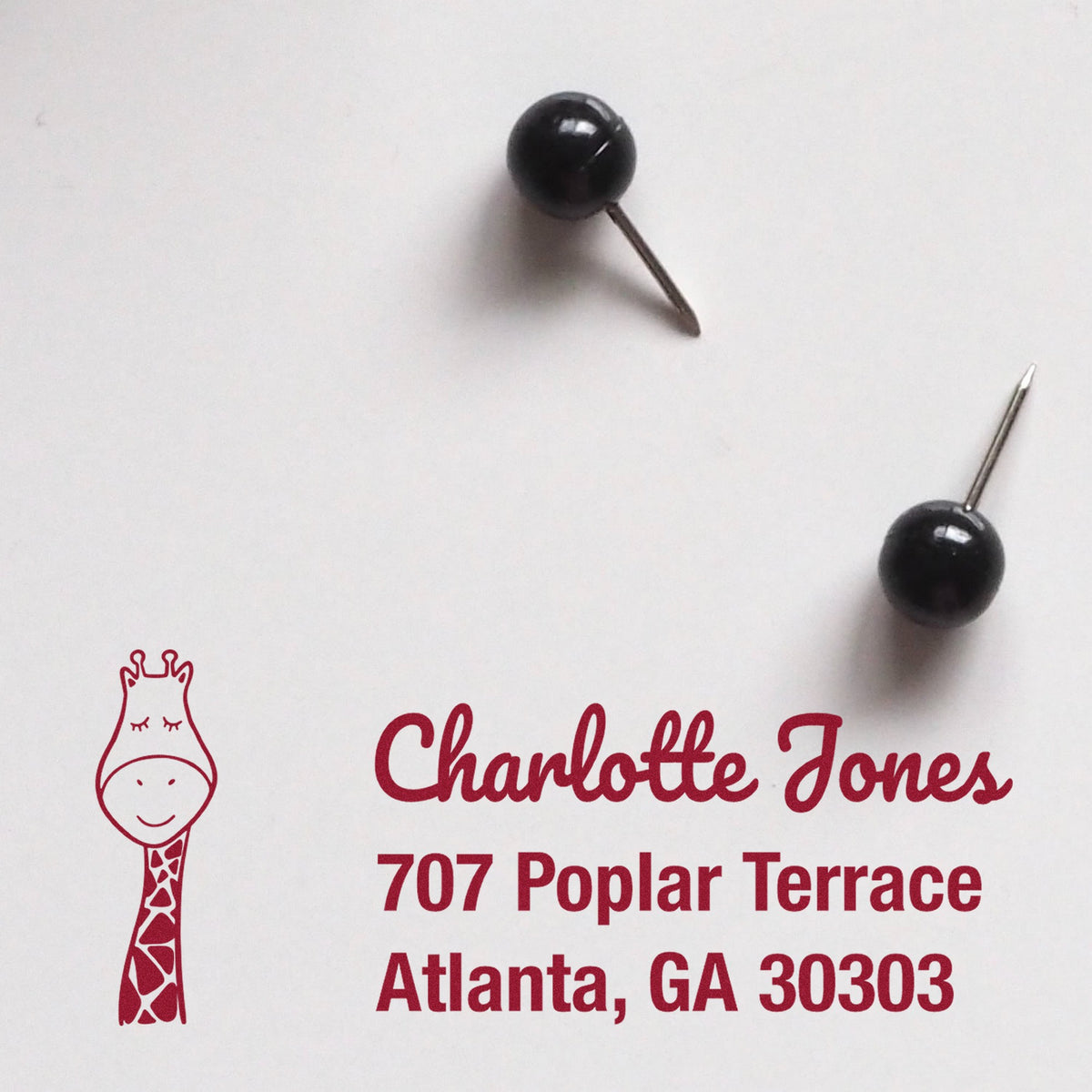 Slim Pre-Inked Jolly Giraffe Custom Address Return Stamp on white paper with text "Charlotte Jones, 707 Poplar Terrace, Atlanta, GA 30303" in red. Two black pushpins nearby. Custom-Made Return Mail Stamper design features a smiling giraffe.