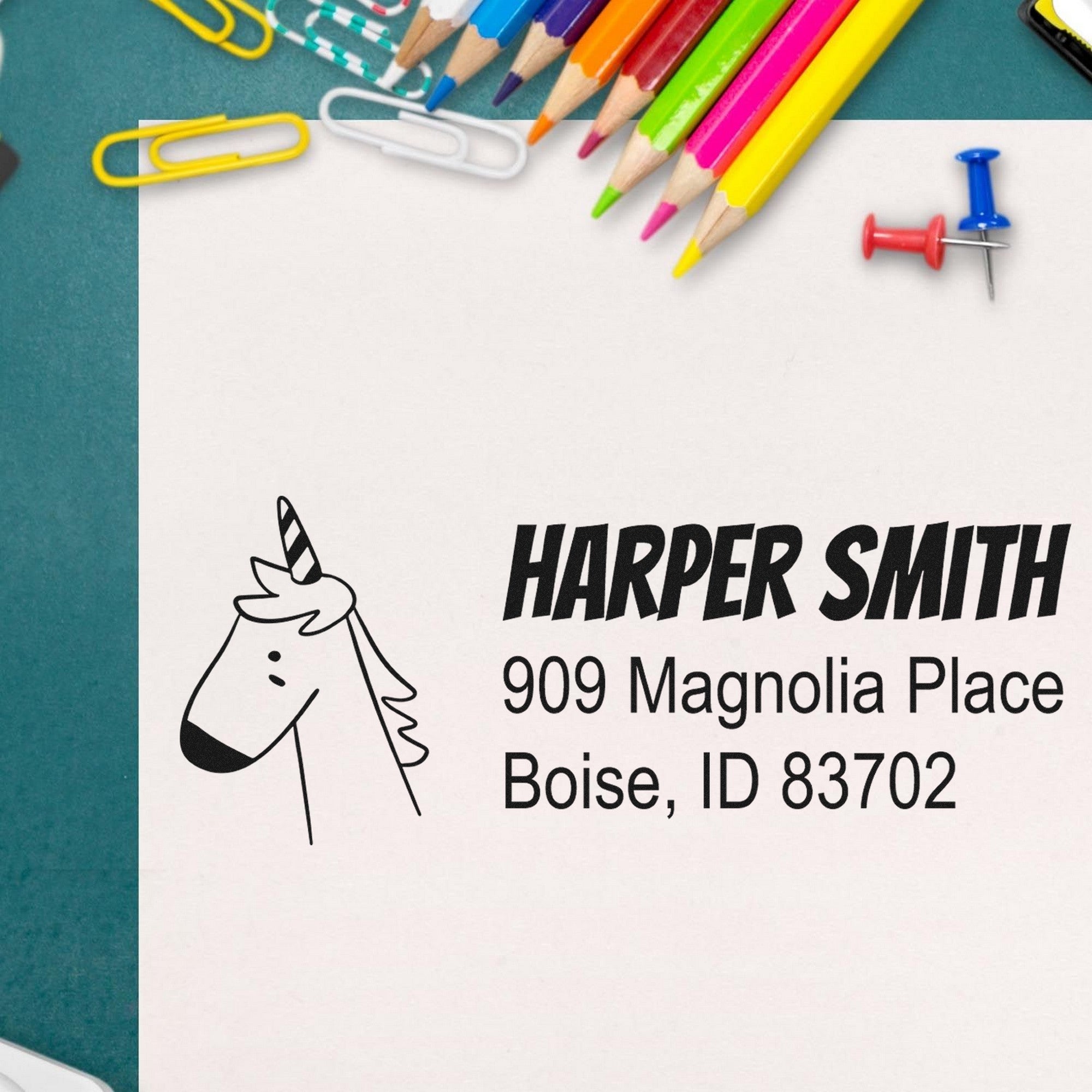 PSI Whimsical Unicorn Handmade Home Address Pre-Inked Stamp on an envelope with "Harper Smith, 909 Magnolia Place, Boise, ID 83702." Surrounded by colorful pencils and paperclips. Personalizable Mailing Stamp for creative correspondence.