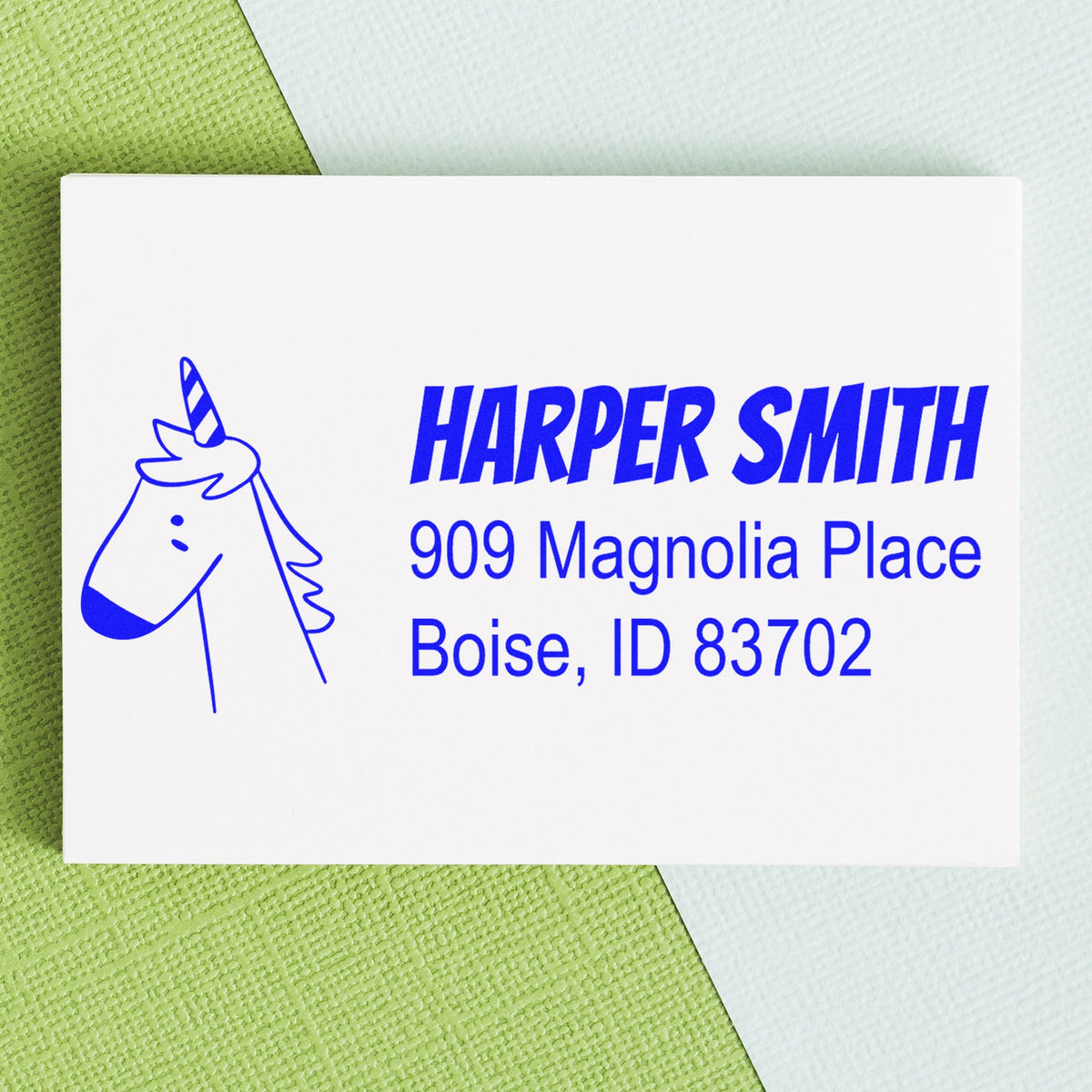 Whimsical Unicorn Customizable Name and Address Self-Inking Stamp featuring a blue unicorn illustration. Personalizable Address Stamp with the name "Harper Smith" and address "909 Magnolia Place, Boise, ID 83702" on a white card against a green and blue background.