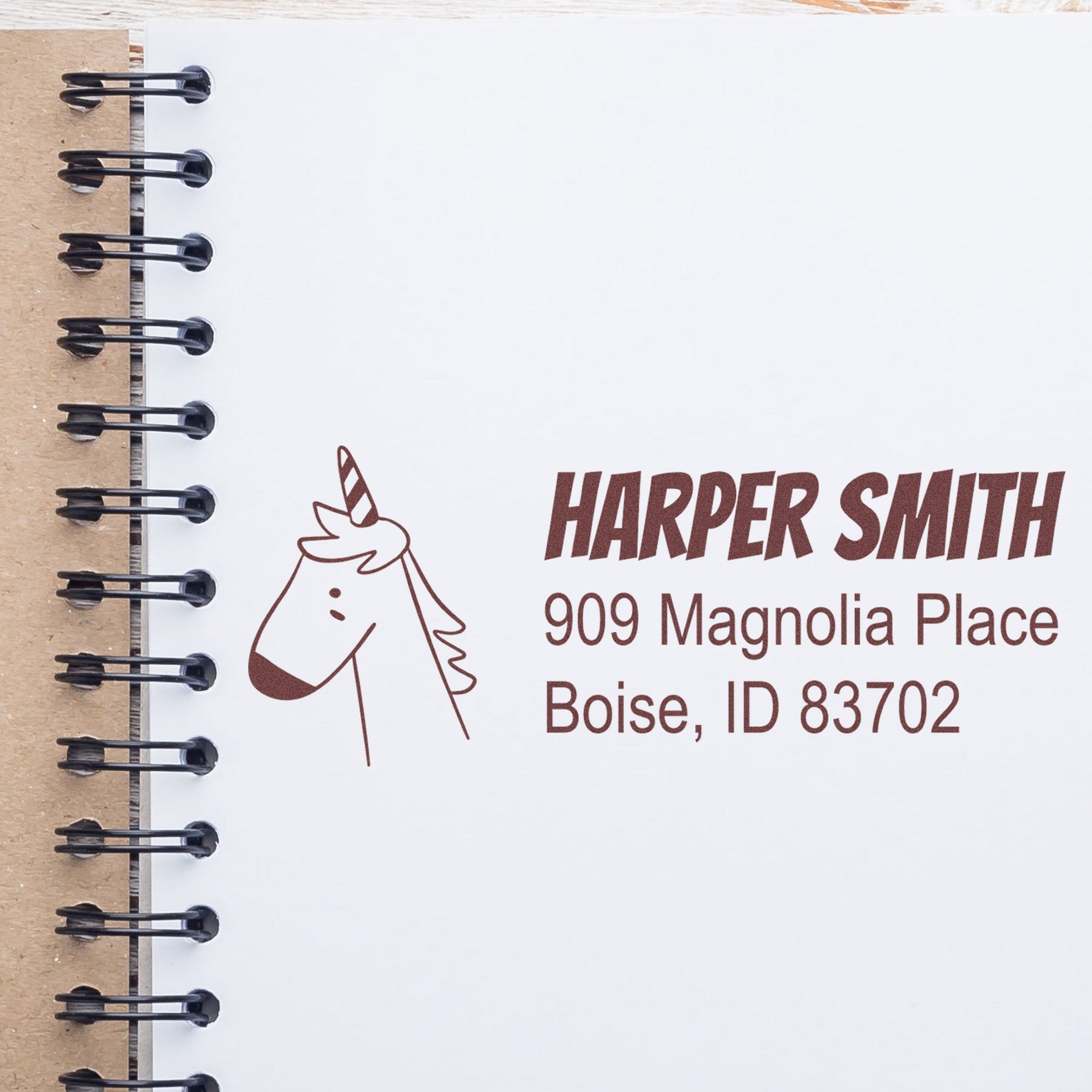 Wood handle whimsical unicorn personalized address label rubber stamp on a notebook page. The stamp features a unicorn design with the text "Harper Smith, 909 Magnolia Place, Boise, ID 83702." Ideal for a personalized mailing stamp.