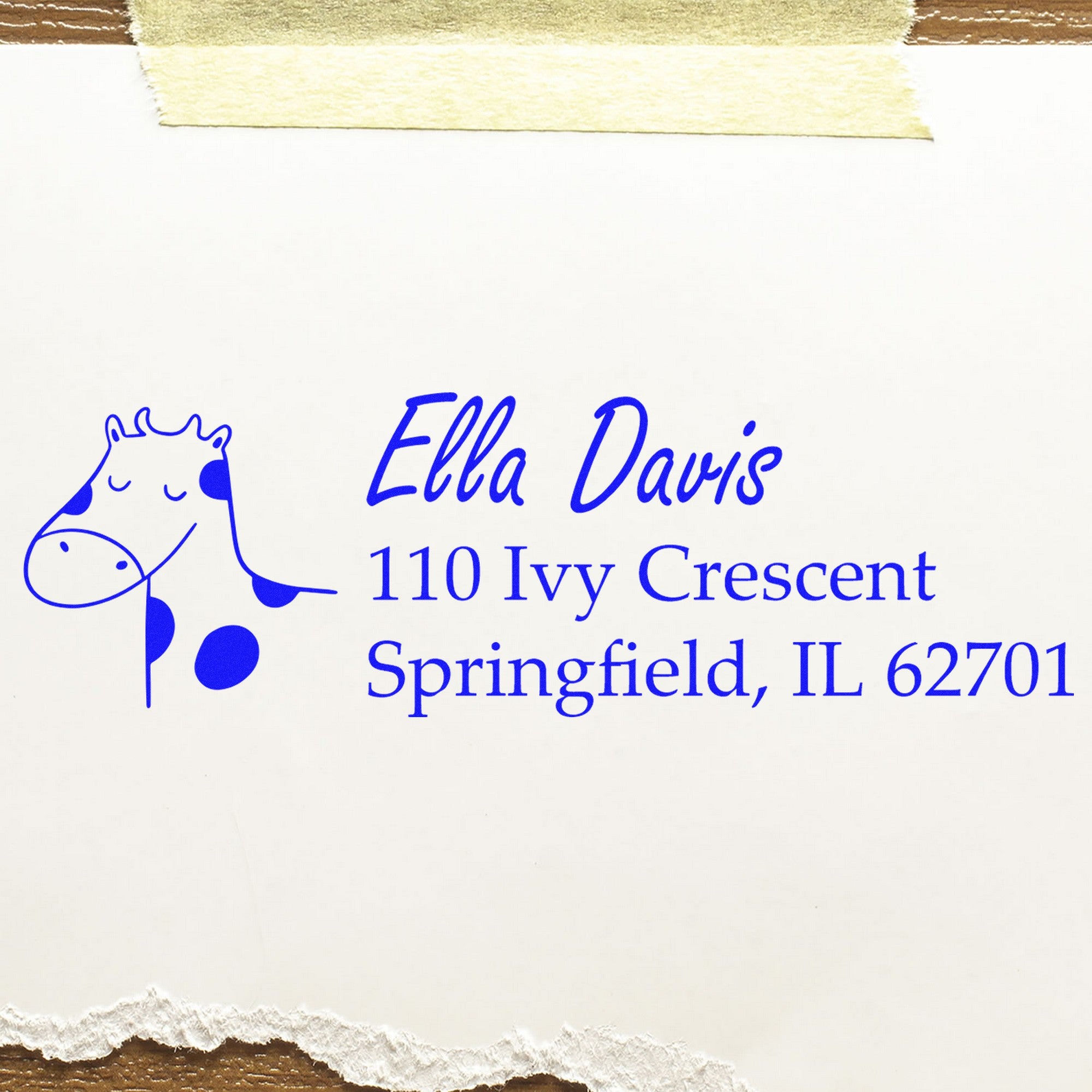 Wood Handle Giraffe Grins Custom Address Rubber Stamp on white paper, featuring a blue giraffe illustration and text: "Ella Davis, 110 Ivy Crescent, Springfield, IL 62701." The Custom Home Stamper is taped to a surface with a torn edge.