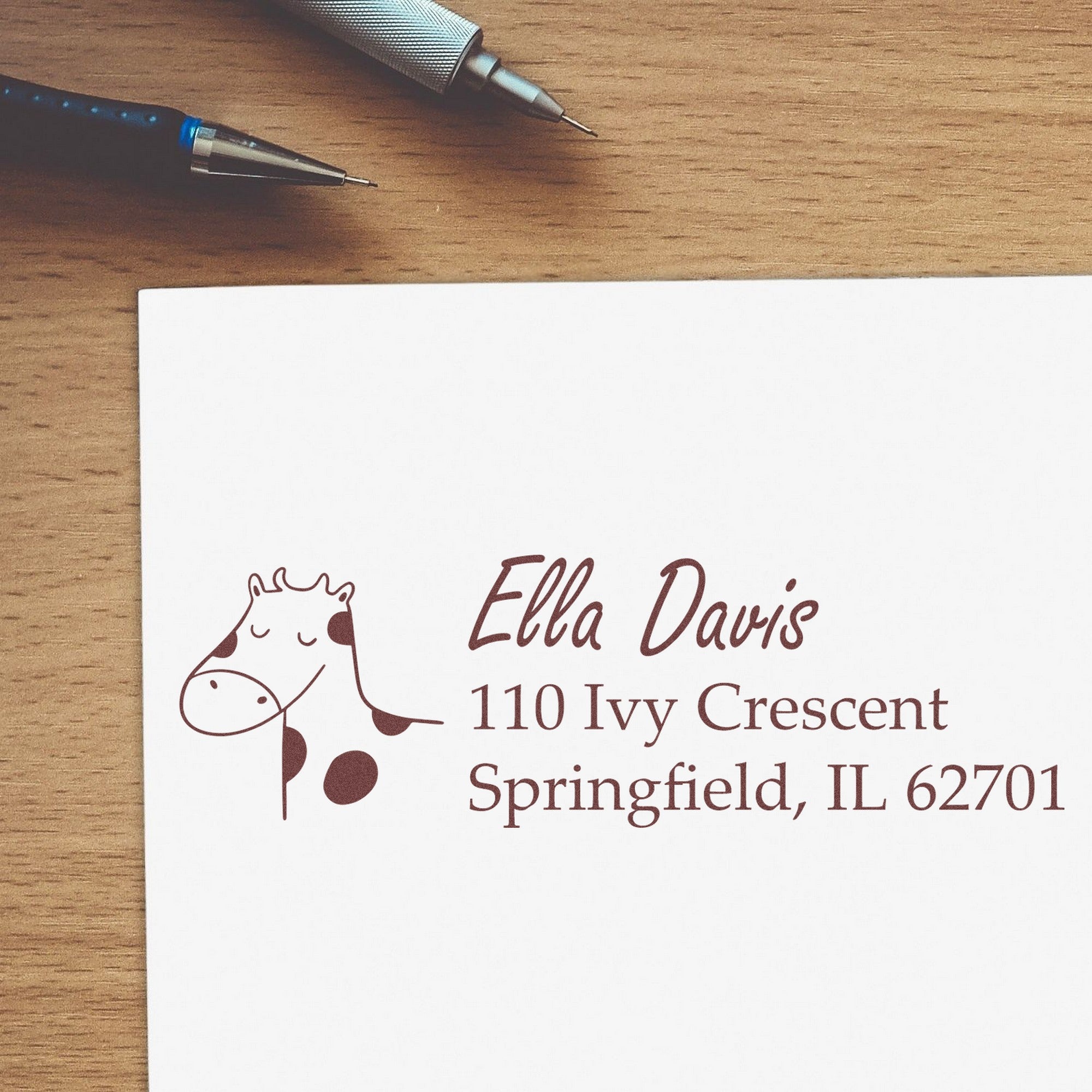 Wood Handle Giraffe Grins Custom Address Rubber Stamp on an envelope with "Ella Davis, 110 Ivy Crescent, Springfield, IL 62701" in brown ink. Custom Return Mail Stamp design features a smiling giraffe illustration, placed on a wooden desk with pens nearby.