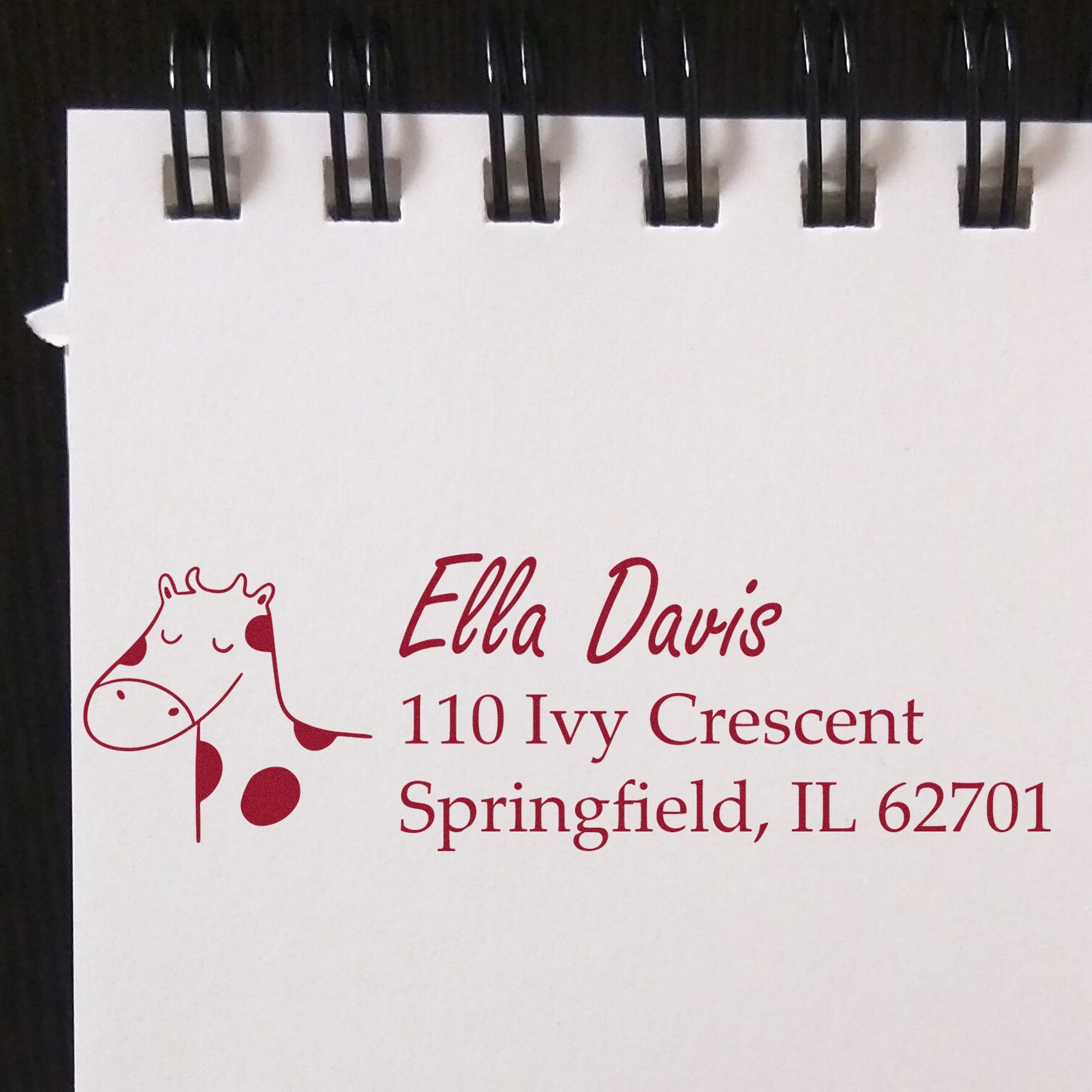 Wood Handle Giraffe Grins Custom Address Rubber Stamp on a notepad, displaying "Ella Davis, 110 Ivy Crescent, Springfield, IL 62701" in red ink. This Custom Mailing Stamper features a smiling giraffe design, ideal for personalized correspondence.