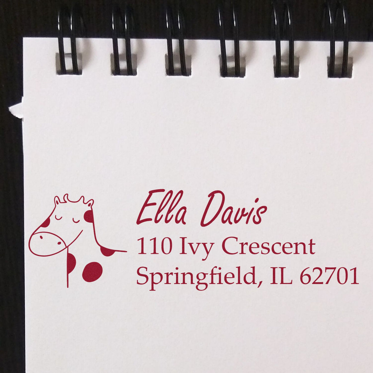 A Slim Pre-Inked Giraffe Grins Custom New Address Stamp is shown on a notepad, featuring a cute giraffe illustration and custom mailing stamp details for Ella Davis, 110 Ivy Crescent, Springfield, IL 62701, in red ink.