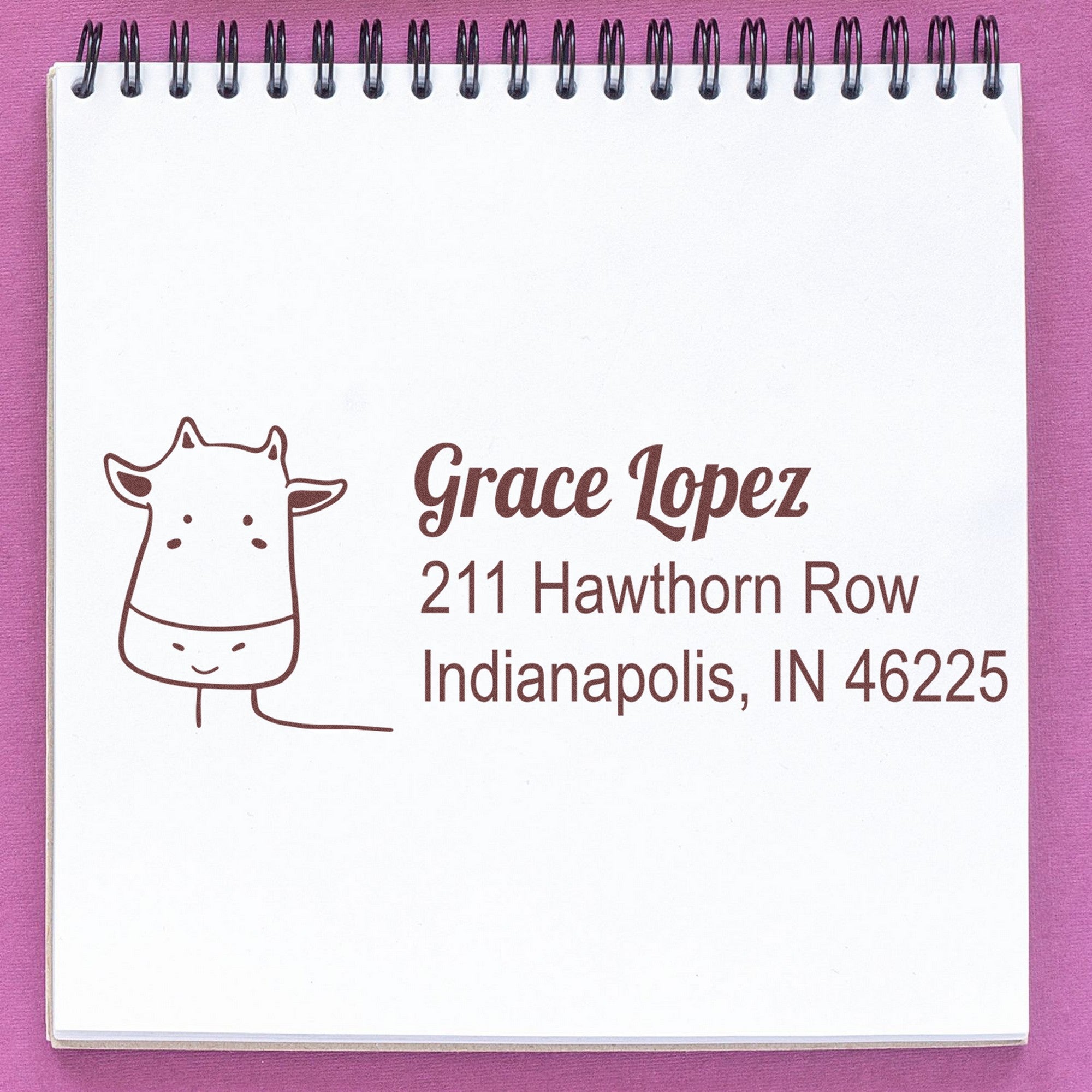 Wood Handle Cow Cuddles Custom Return Address Rubber Stamp on a notepad, featuring a cow illustration and the text "Grace Lopez, 211 Hawthorn Row, Indianapolis, IN 46225." This Customizable New Address Stamp is displayed on a spiral-bound notebook.