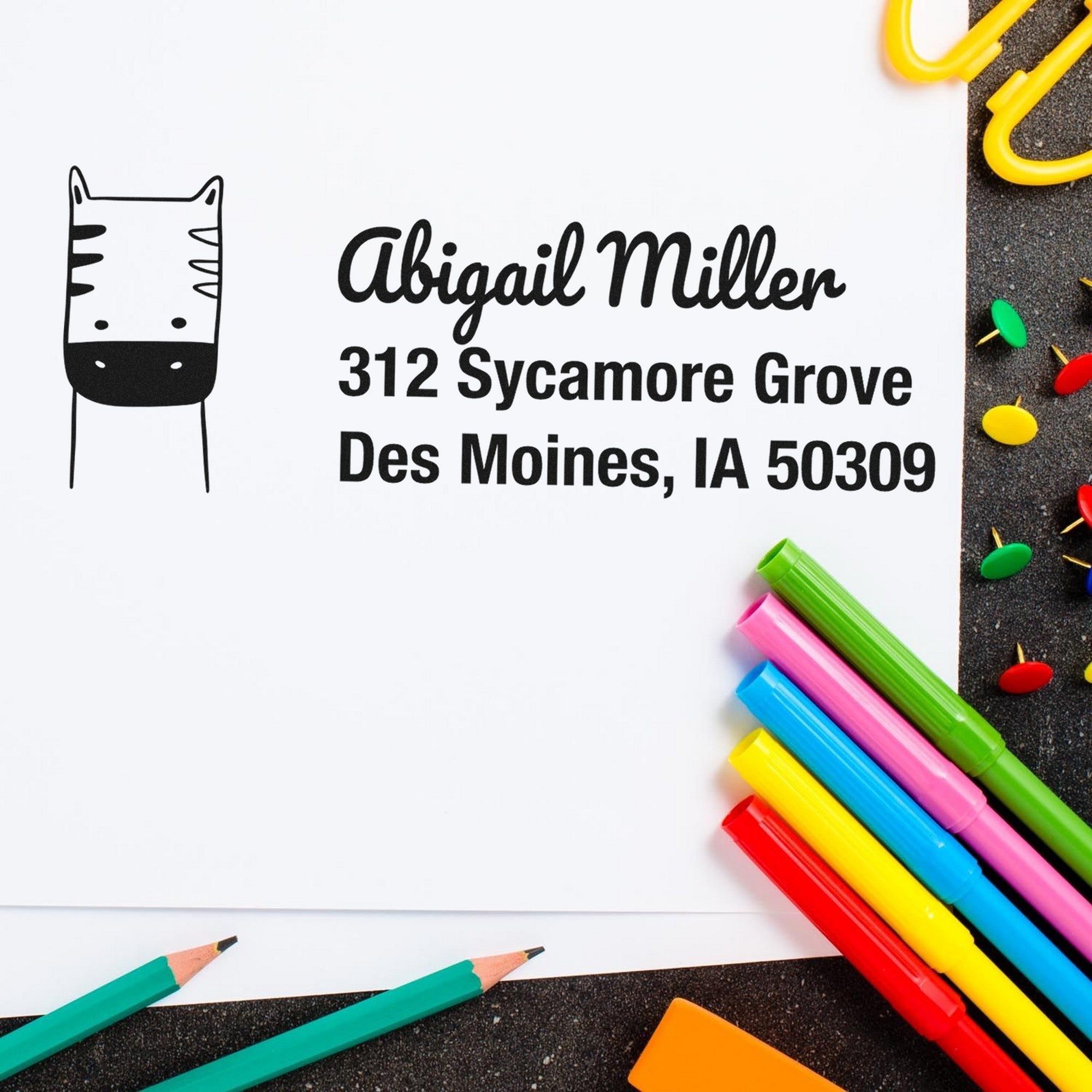 PSI Frolicsome Zebra Handmade Mail Address Pre-Inked Stamp on white paper with "Abigail Miller, 312 Sycamore Grove, Des Moines, IA 50309" text. Custom-Made Home Stamper surrounded by colorful pens and paperclips on a dark surface.
