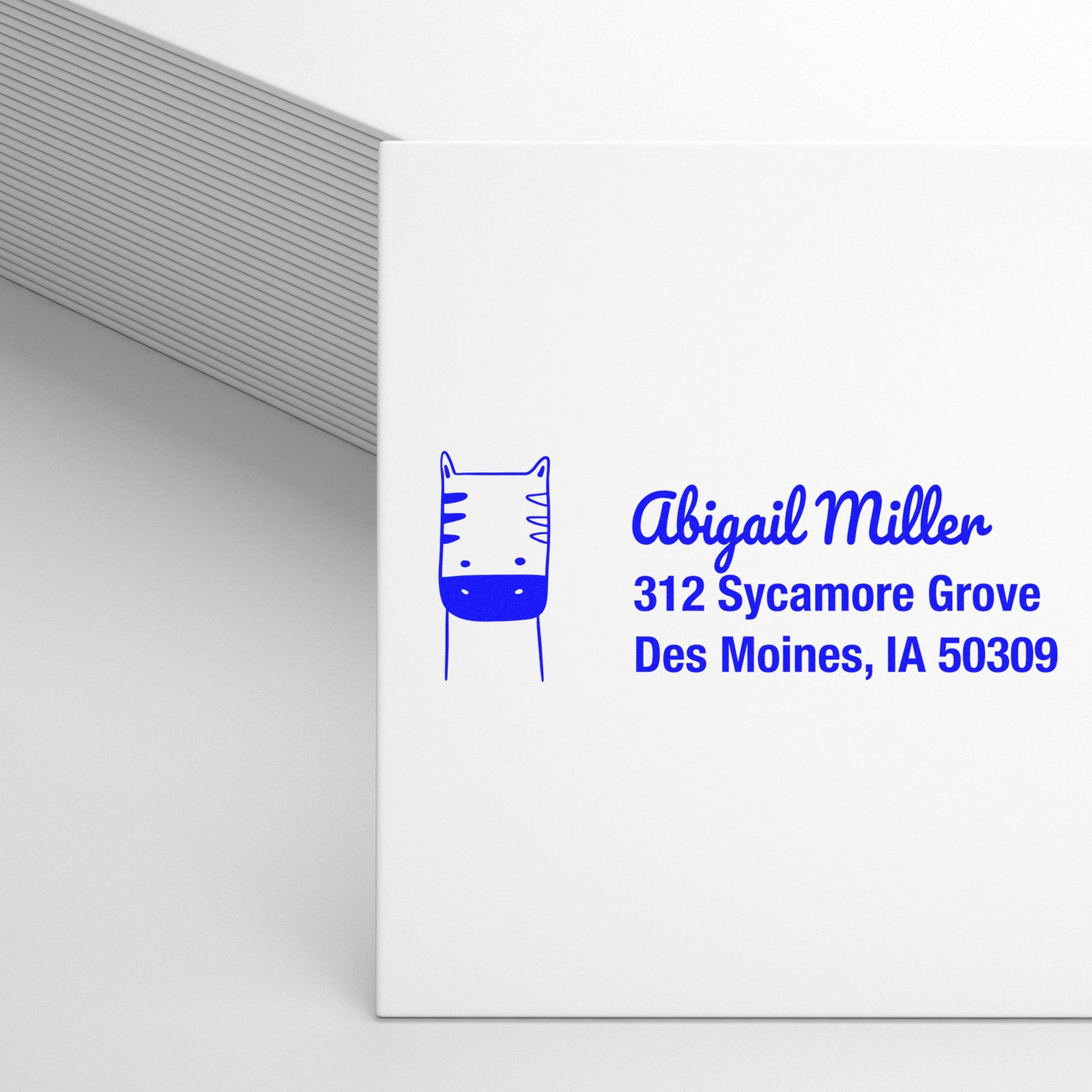 Image of a white card featuring a blue frolicsome zebra design with a custom name and address. The text reads "Abigail Miller, 312 Sycamore Grove, Des Moines, IA 50309." A wood handle rubber stamp is nearby, showcasing a personalizable new address stamper.
